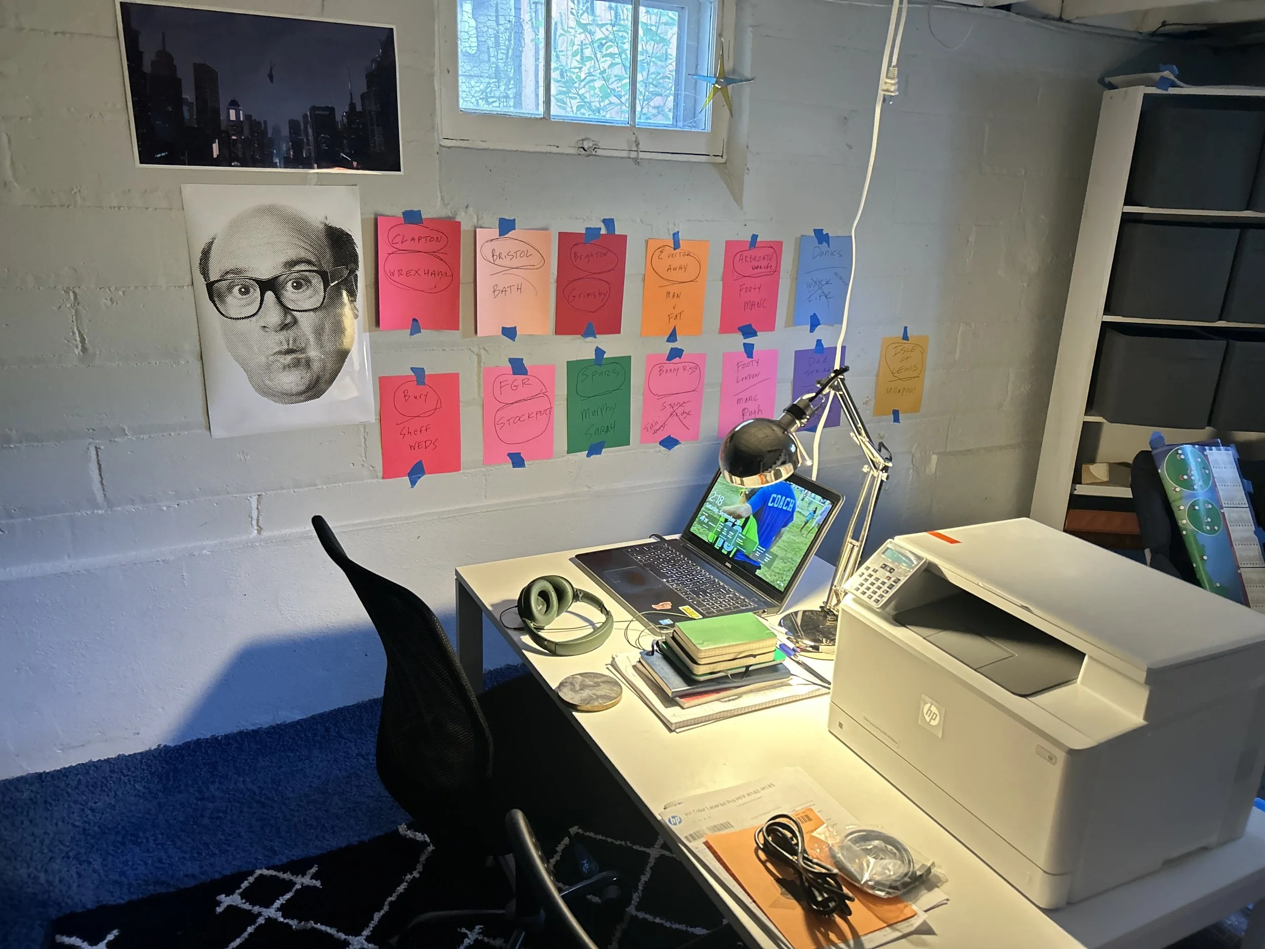 An office workspace with a white desk holding a laptop, a printer, a lamp, headphones, notebooks, and a few other items. Behind the desk, a wall features colorful notes taped with blue tape and a large black and white photo of a man with glasses and 