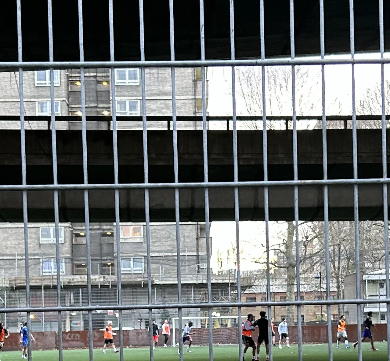 Soccer players practicing or playing on a field viewed through a metal fence. A building and trees are visible in the background.