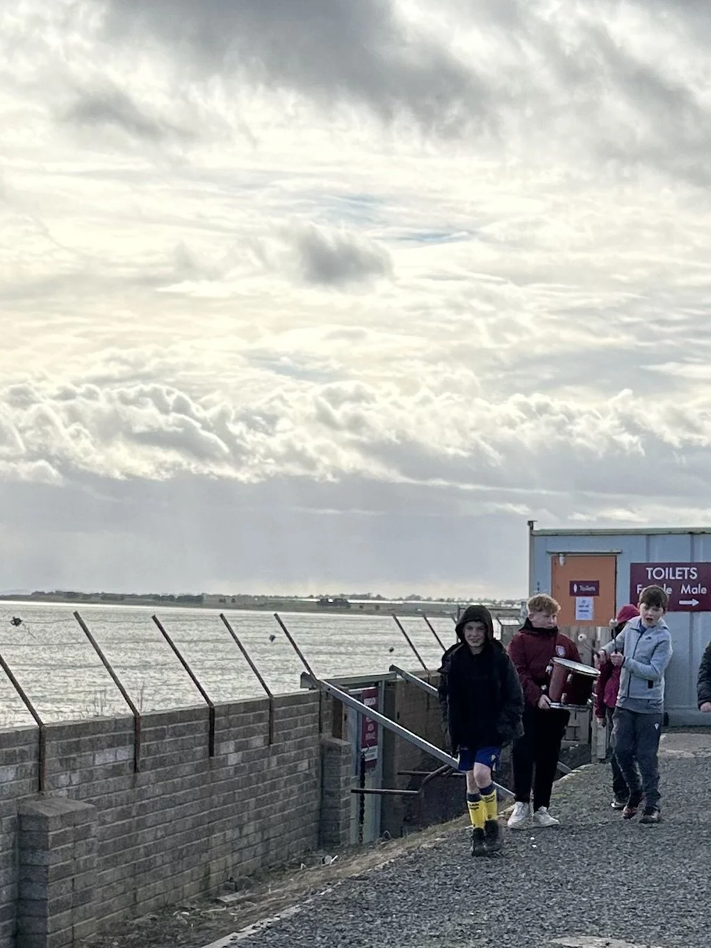 Group of young boys walking near the water on a cloudy day, with a building labeled "Toilets" in the background.