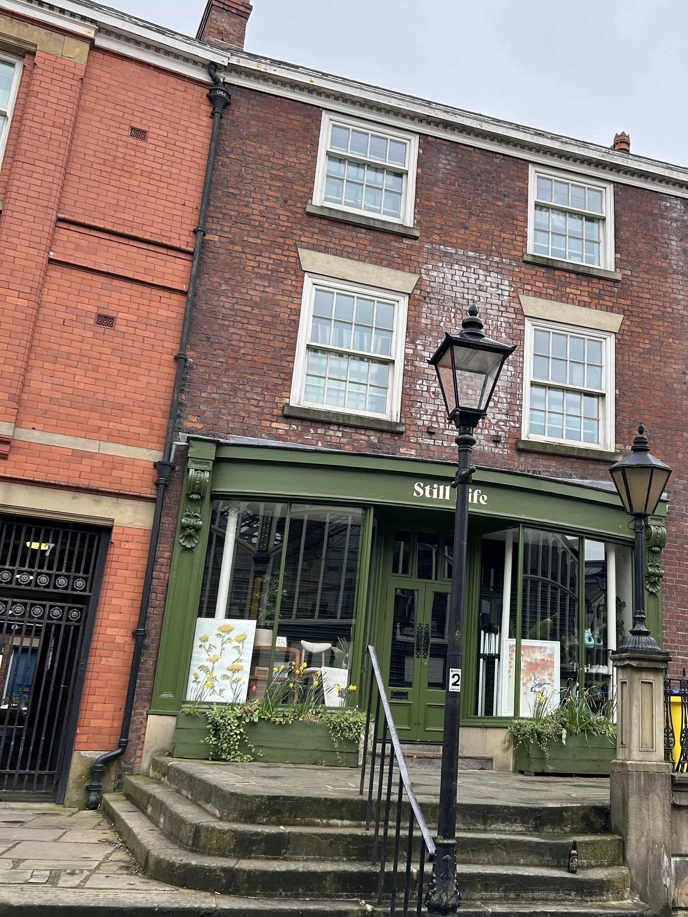 A brick building with a green storefront named 'Still Life' with large windows displaying plants and artwork. There are two black street lamps in front, and steps leading up to the entrance.