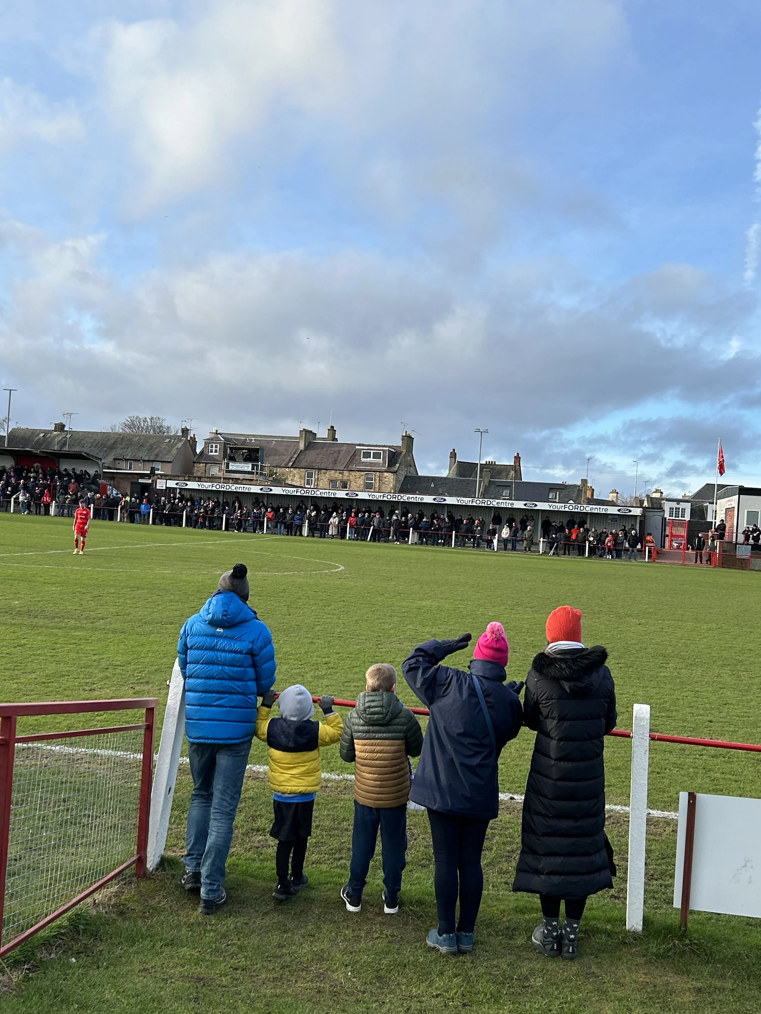 Spectators watching a football game from the sidelines at a small stadium, with players on the field and houses in the background under a cloudy sky.