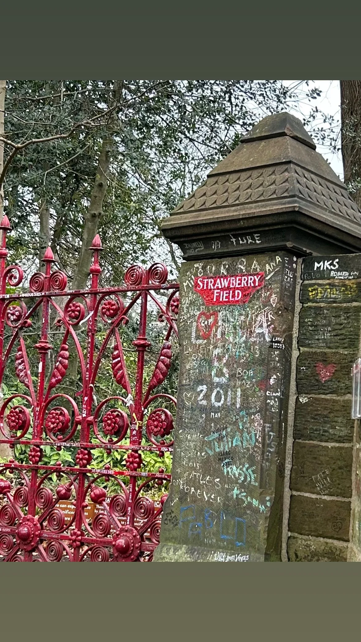 A stone entrance pillar with graffiti and a red metal gate pointing to the Strawberry Field attraction, with trees in the background.