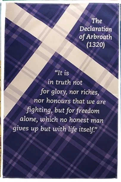A plaid-patterned book or poster with a quote from the Declaration of Arbroath (1320). The quote emphasizes the importance of freedom over fame, riches, or honors, highlighting that honest men give up everything but retain life itself.