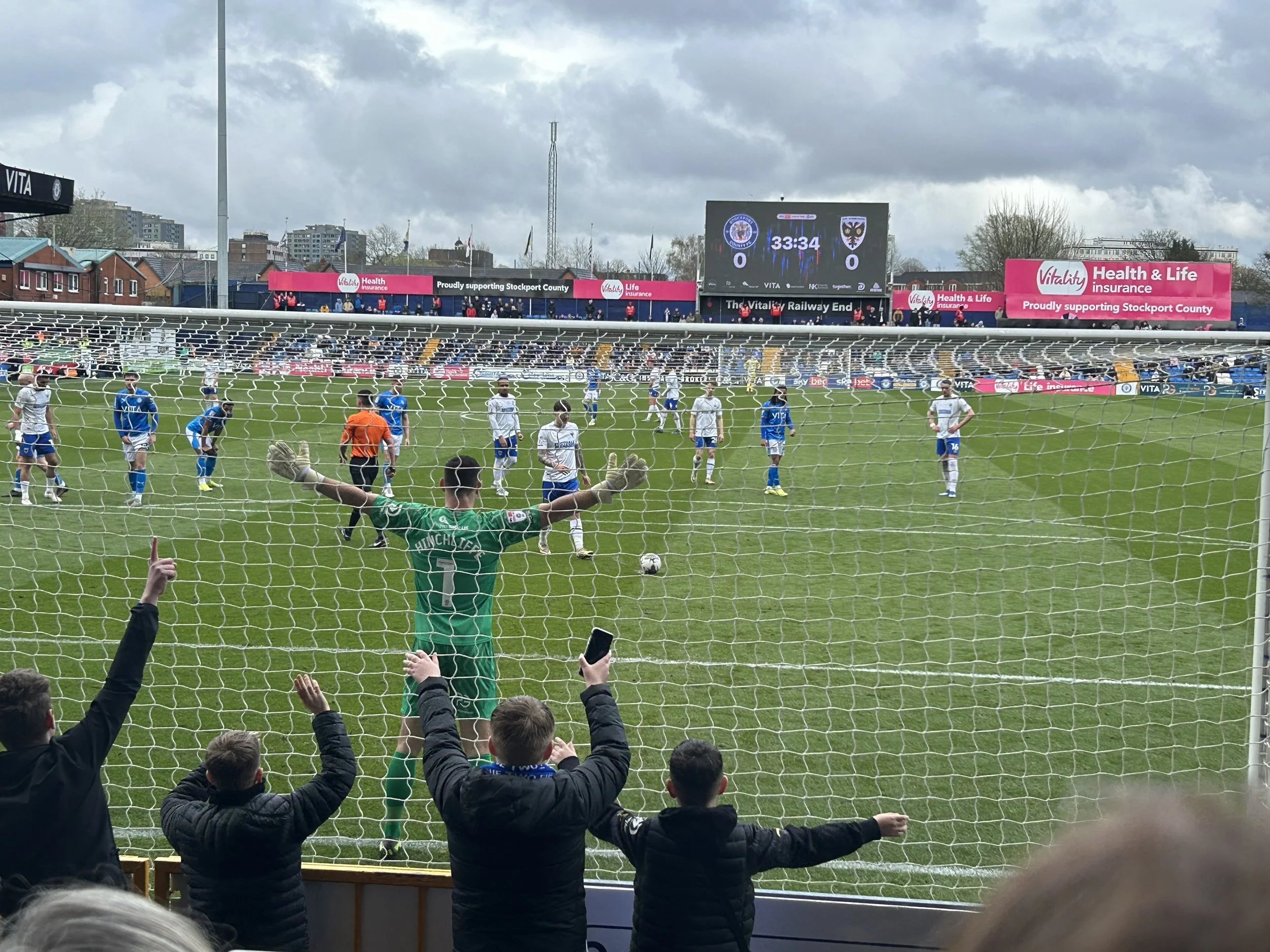 View of a soccer field during a match with players on the field, fans in the stands, and a large scoreboard displaying 33 minutes and 34 seconds with scores tied at 0-0.