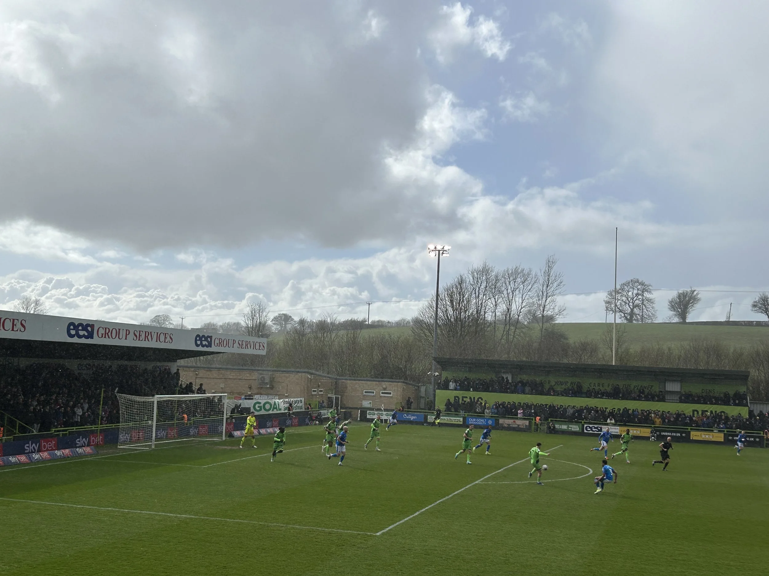 A soccer match in progress with players on the field, a goalpost, and spectators in the stands under a partly cloudy sky.