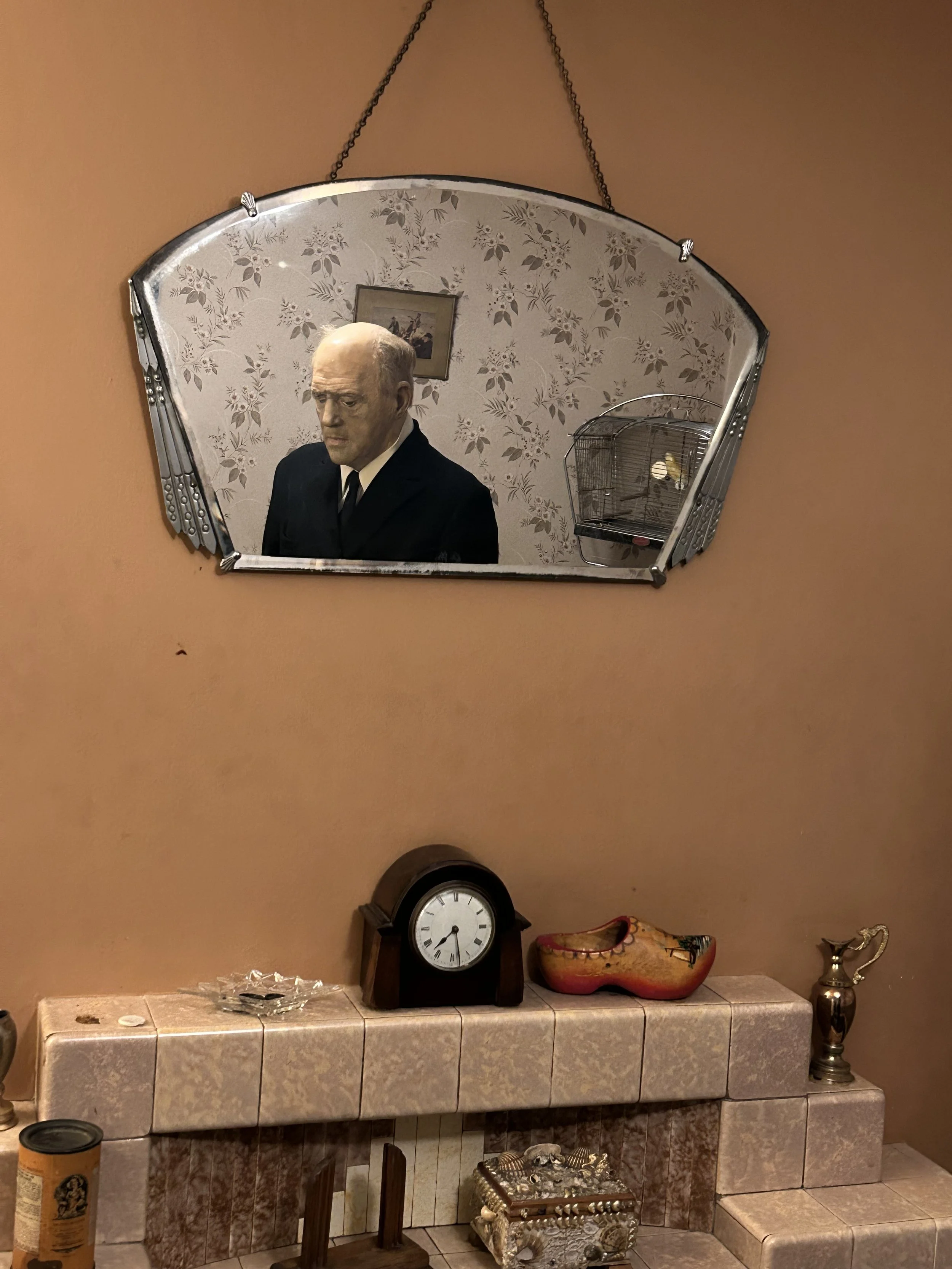 A mirror on the wall reflects a mannequin head of an elderly man with a serious expression, wearing a black suit. The room behind the mirror has floral wallpaper and a picture on the wall. Below, there is a tiled fireplace with various decorative ite