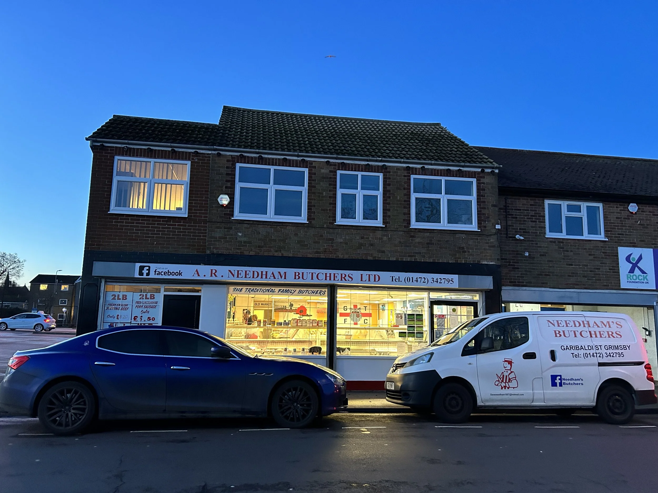 Street view of A. R. Needham Butchers shop with two cars parked outside, including a white delivery van, on a clear evening with a blue sky. The shop has windows displaying meat products and signage promoting traditional family butchers.