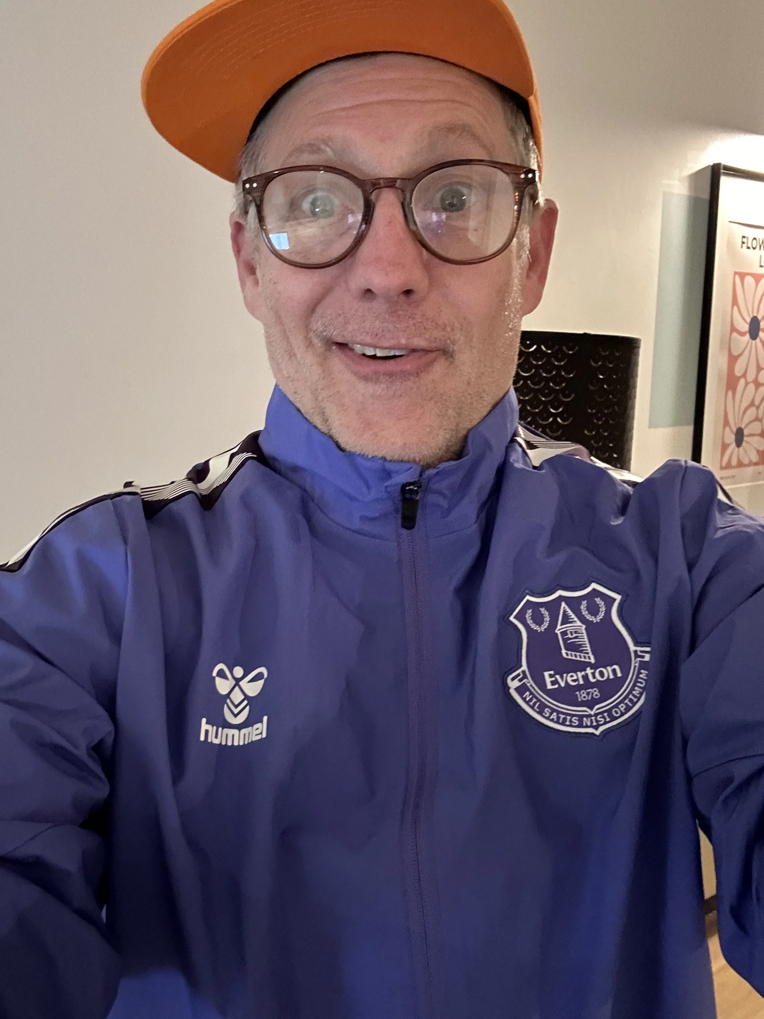 Author Todd Smith taking a selfie, wearing glasses, an orange cap, and a blue sports jacket with the Everton Football Club logo.