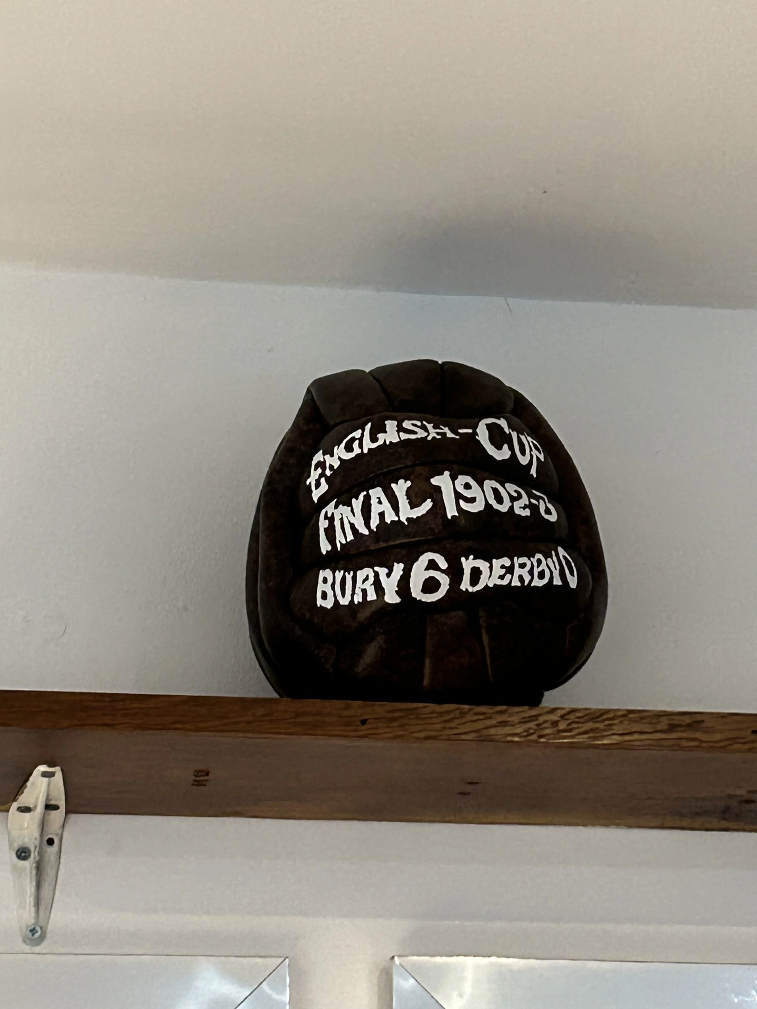 A vintage black soccer ball with white painted text commemorating a final game, resting on a wooden shelf. The text reads: "English Cup Final 1902 - Bury 6 Derby."