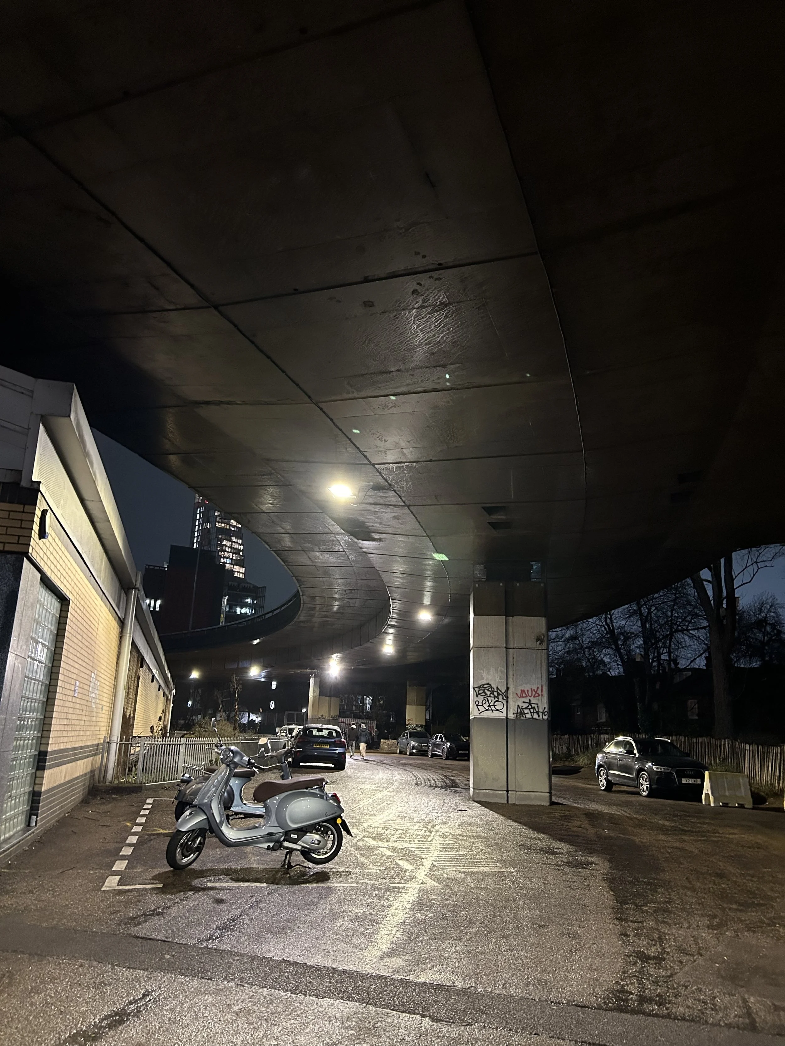 Nighttime scene of a parking area under a curved elevated highway, with a silver scooter parked in the foreground and several cars parked further back, some graffiti on a concrete pillar, and a city skyline with tall buildings in the background.