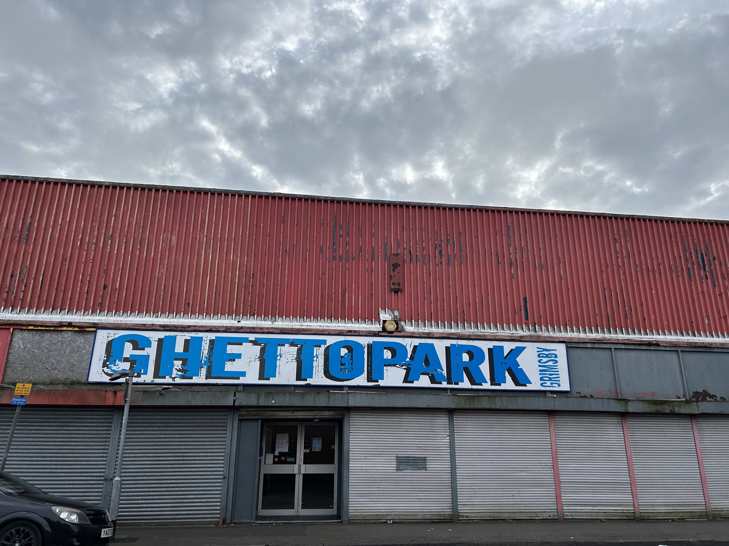 An abandoned building with a blue and white sign that reads 'GHEttO PARK' with graffiti scrapes on it, a metal shutter door, parked cars, and a cloudy sky above.