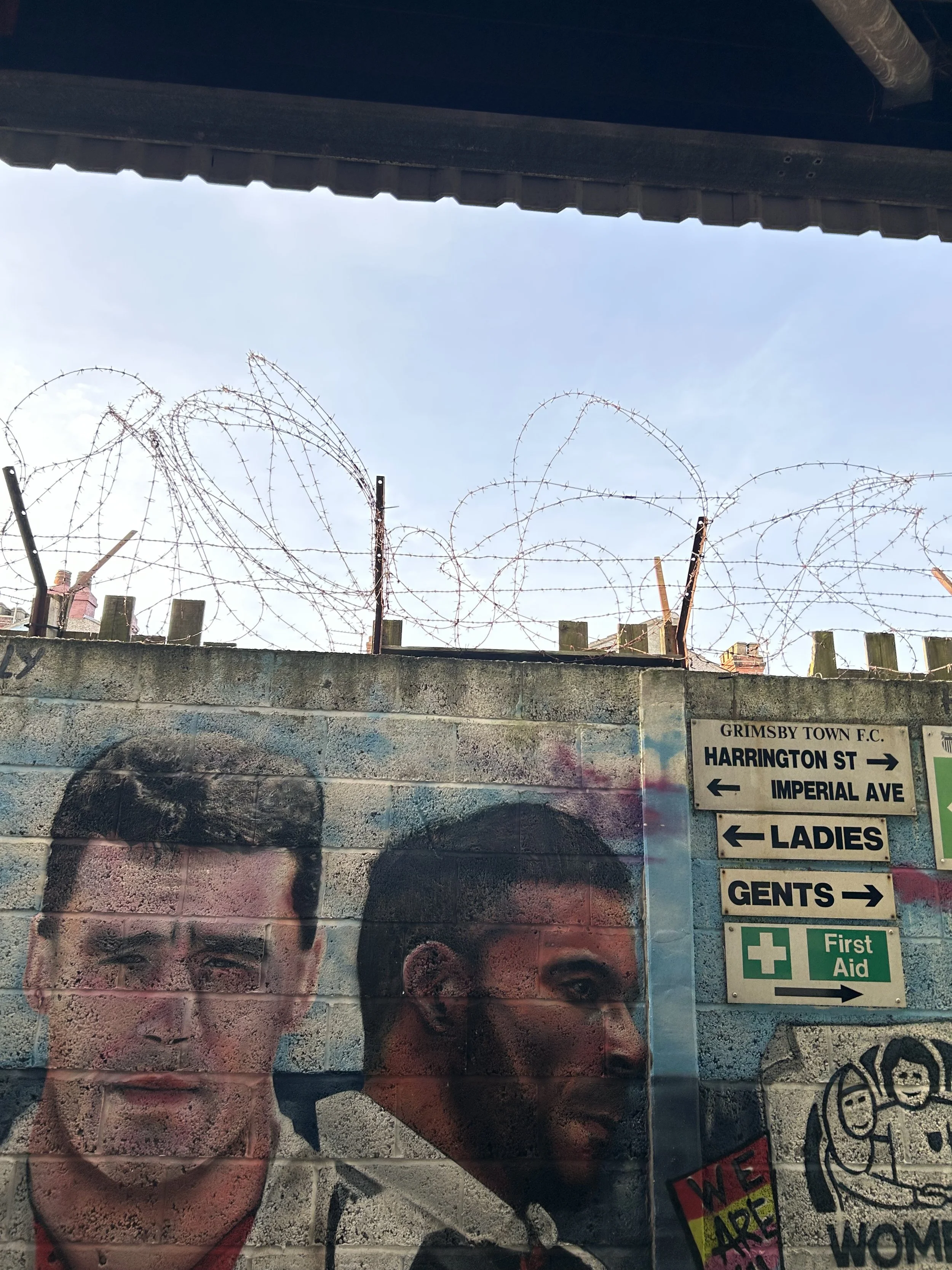Wall with graffiti of two men's portraits and directional signs for Grimsby Town F.C., Harrington Street, Imperial Avenue, Ladies, Gents, and First Aid, barbed wire on top of the wall, and clear sky background.