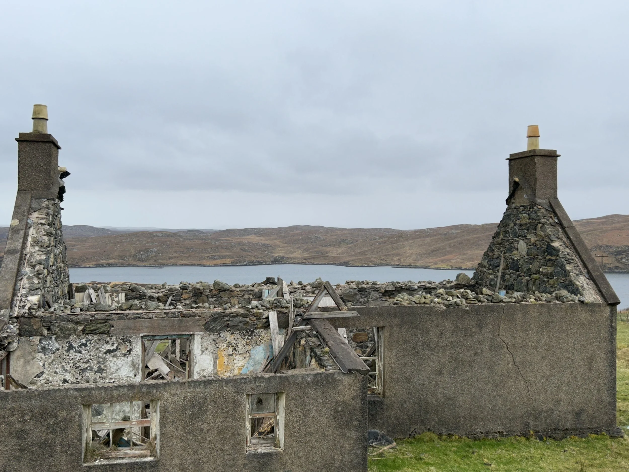 Ruined house with two chimneys, missing roof and broken windows, next to a lake or river with hills in the background.