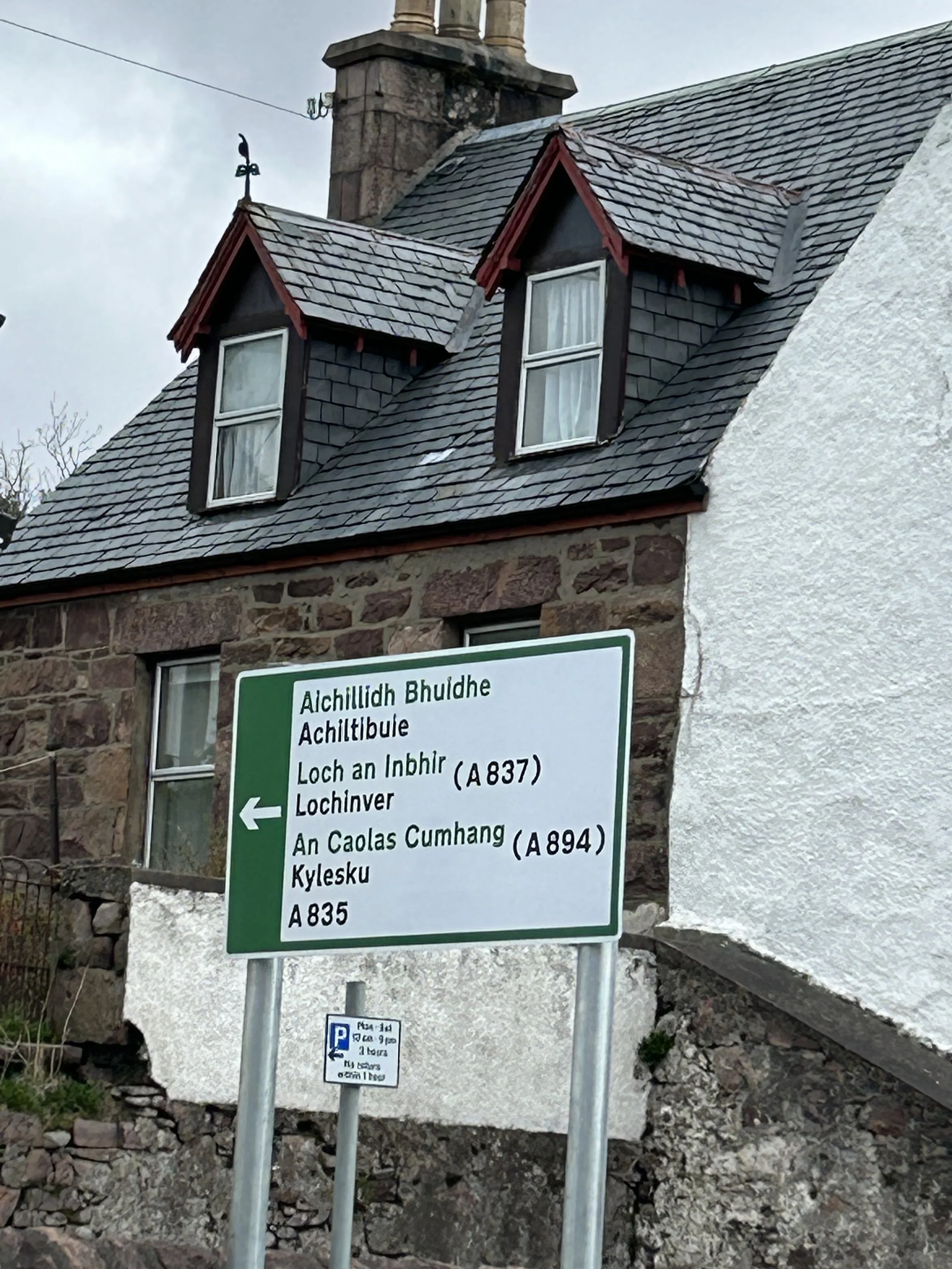 Street sign in front of a stone house with dormer windows. The sign has text in Gaelic and English indicating directions to Achill Island, Loch an Inbhir, An Caolas Cumbang, and Kylesku.