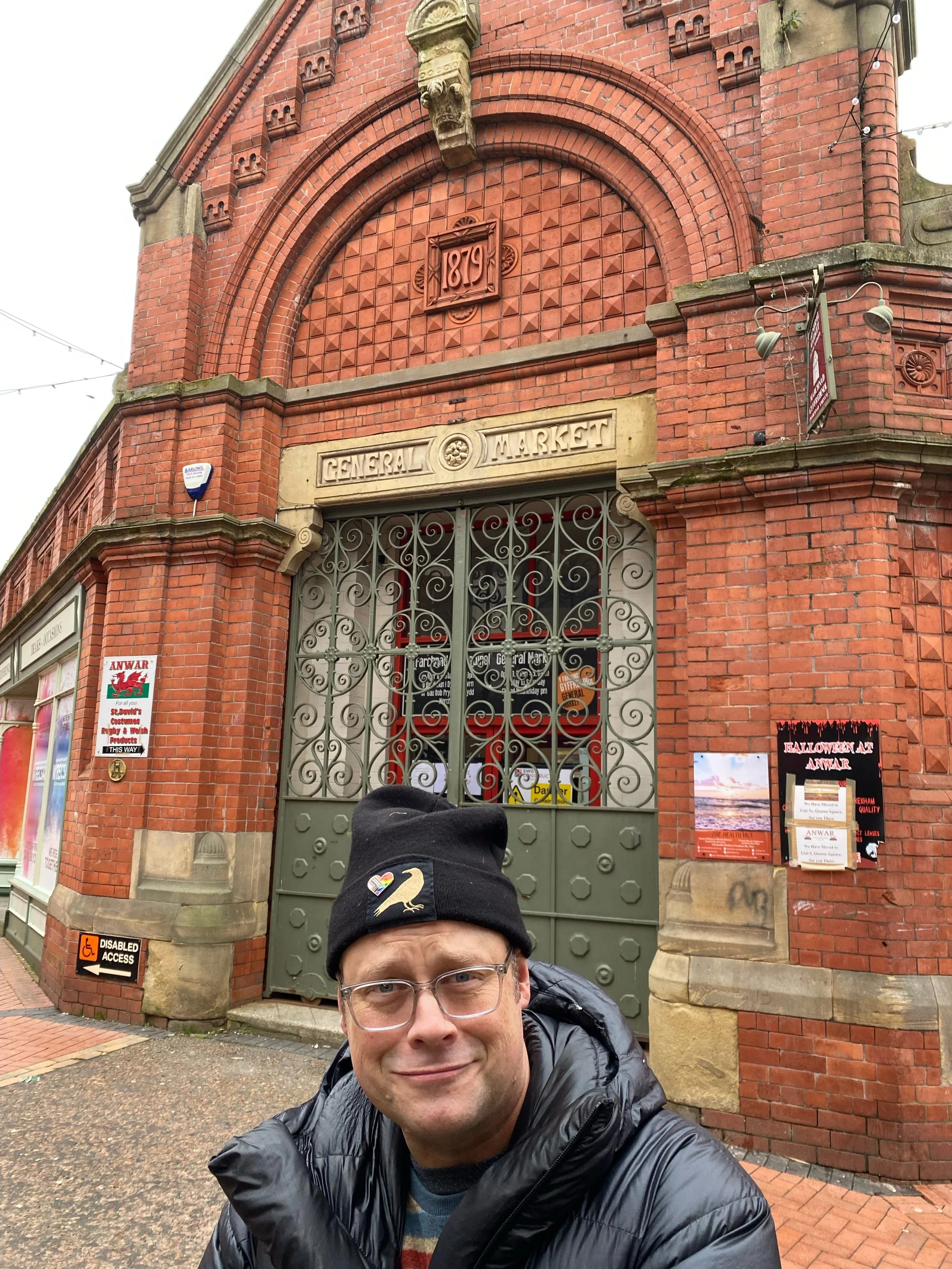 A man wearing glasses, a black beanie with a rainbow swan logo, and a black puffer jacket stands in front of a historic red brick building with an arched gate labeled 'General Market' and the date '1879' inscribed above. The building features decorat