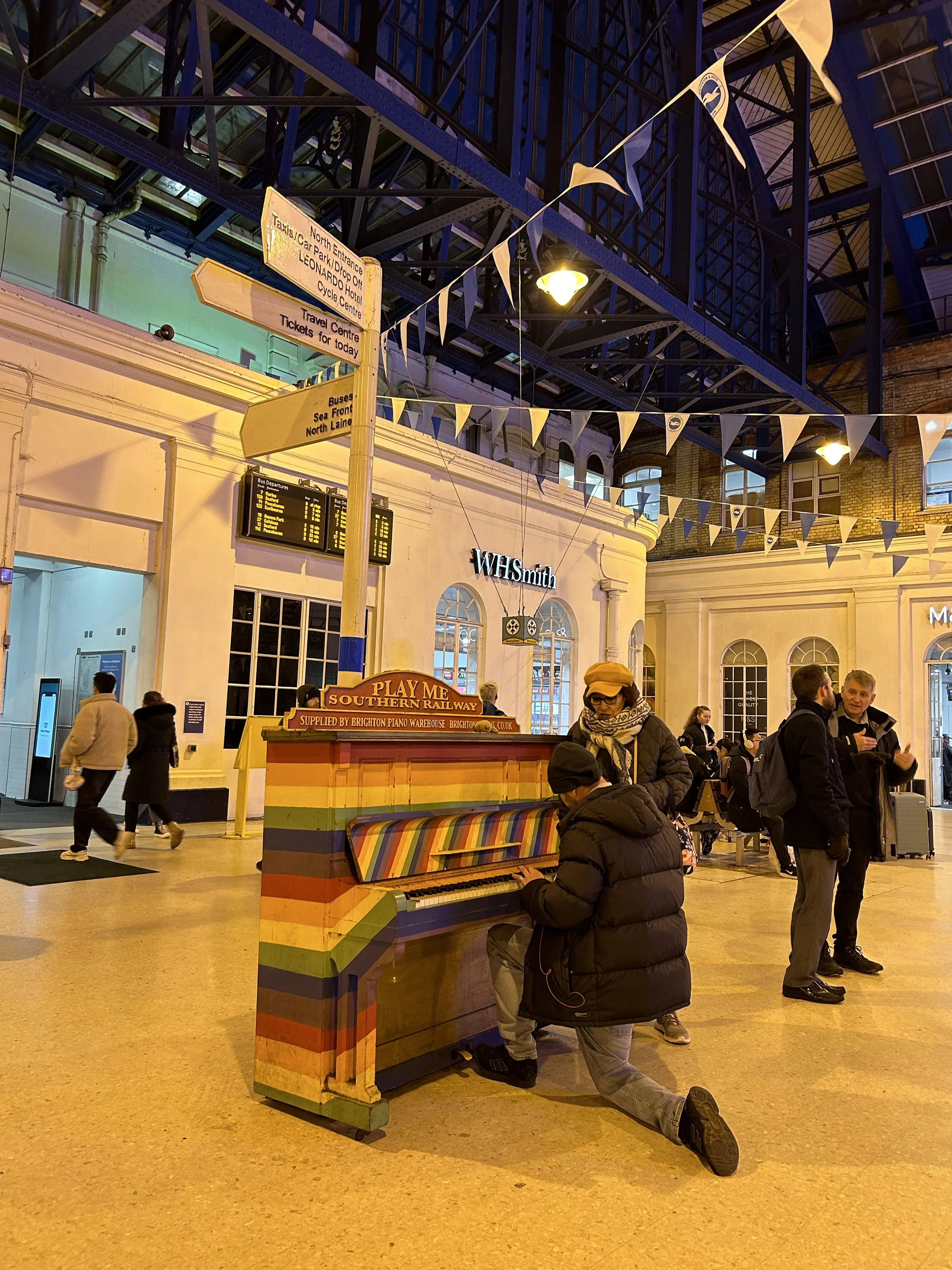 People at a train station with a colorful painted piano in the foreground. The station has high ceilings, large windows, and decorative bunting. A person is playing the piano while others stand nearby, some talking and some walking.