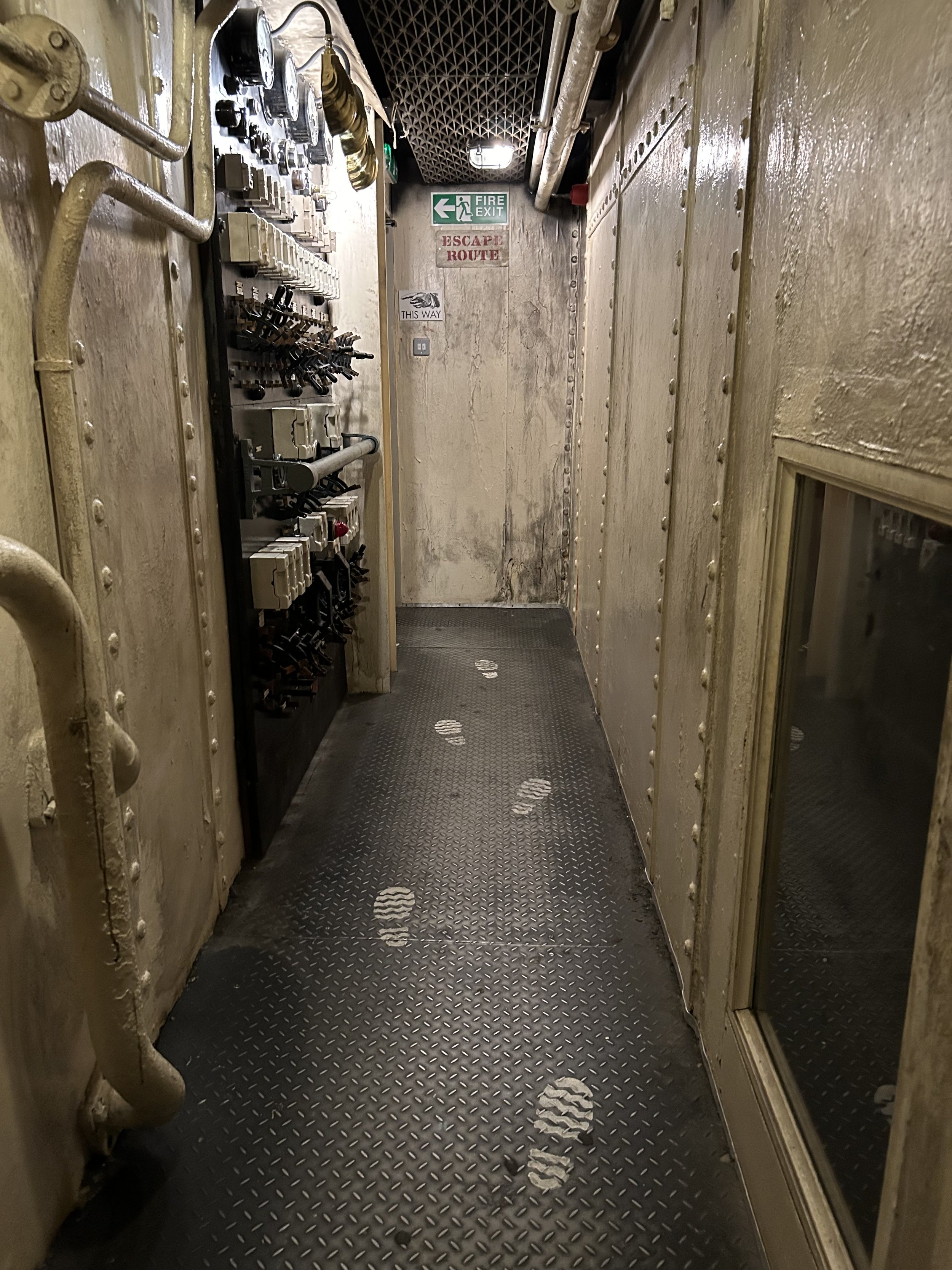 Narrow industrial corridor with footprints on black metal floor, control panels and pipes on the left wall, and a window on the right wall, with signs for fire exit and escape route at the end.