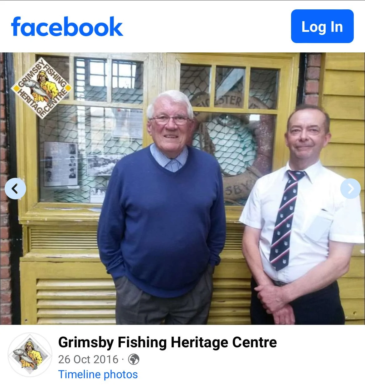 Two men standing in front of a yellow display window at the Grimsby Fishing Heritage Centre. One man is wearing a blue sweater and glasses, and the other is dressed in a white shirt with a patterned tie. The display appears to be related to fishing a