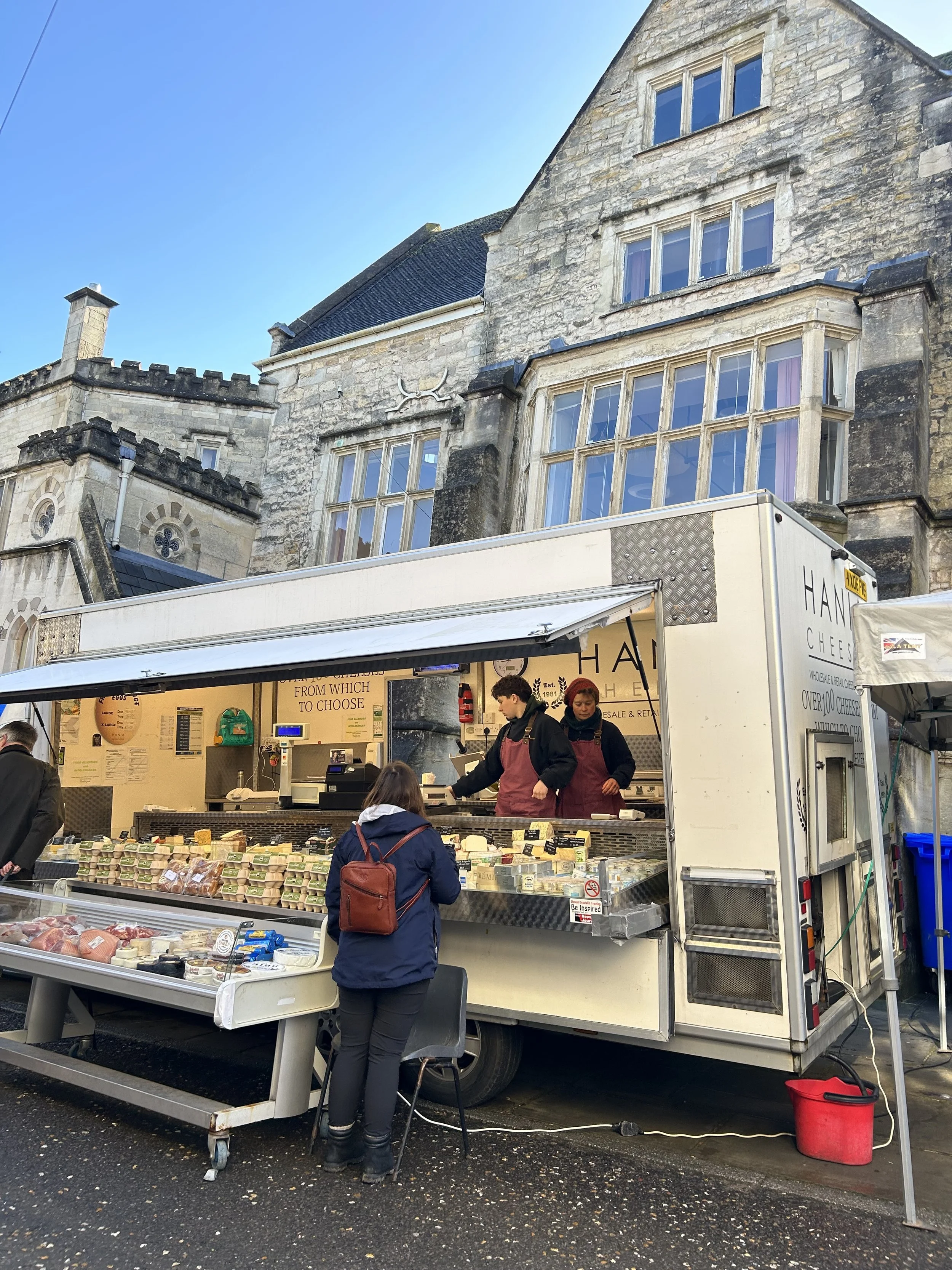 A street market stall selling cheese, with a customer and vendors in front of a historic stone building with large windows.
