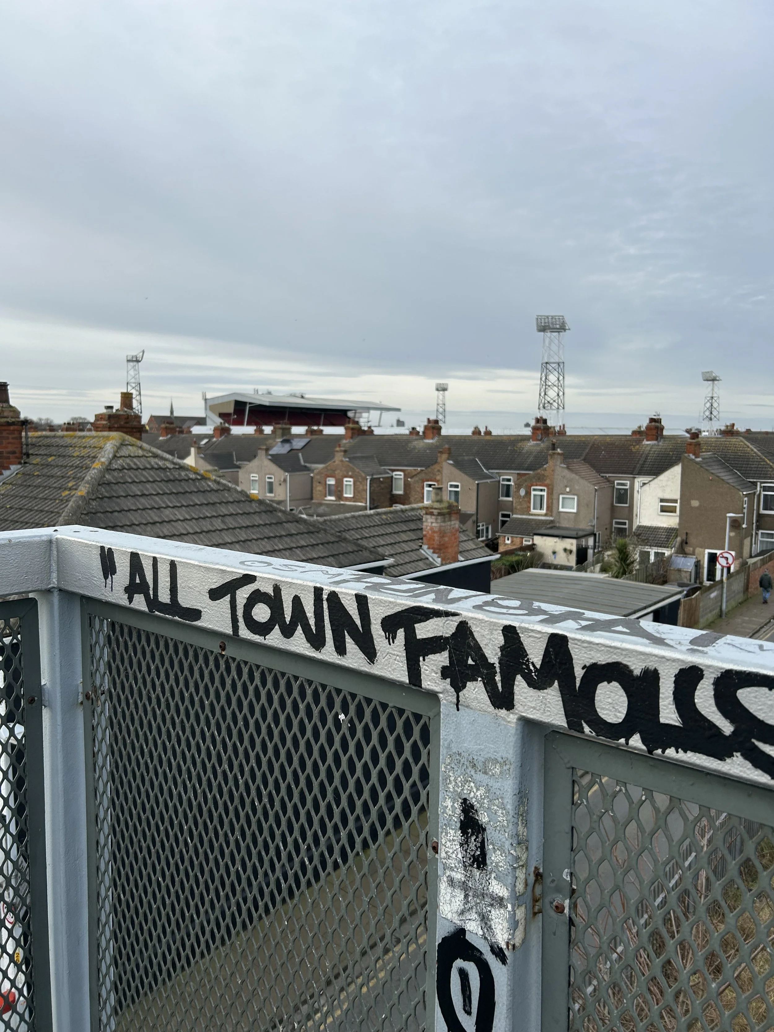 View from a rooftop balcony showing a row of houses, rooftops and cell towers in the background, with graffiti on the balcony railing reading 'ALL TOWN FAMOUS'.