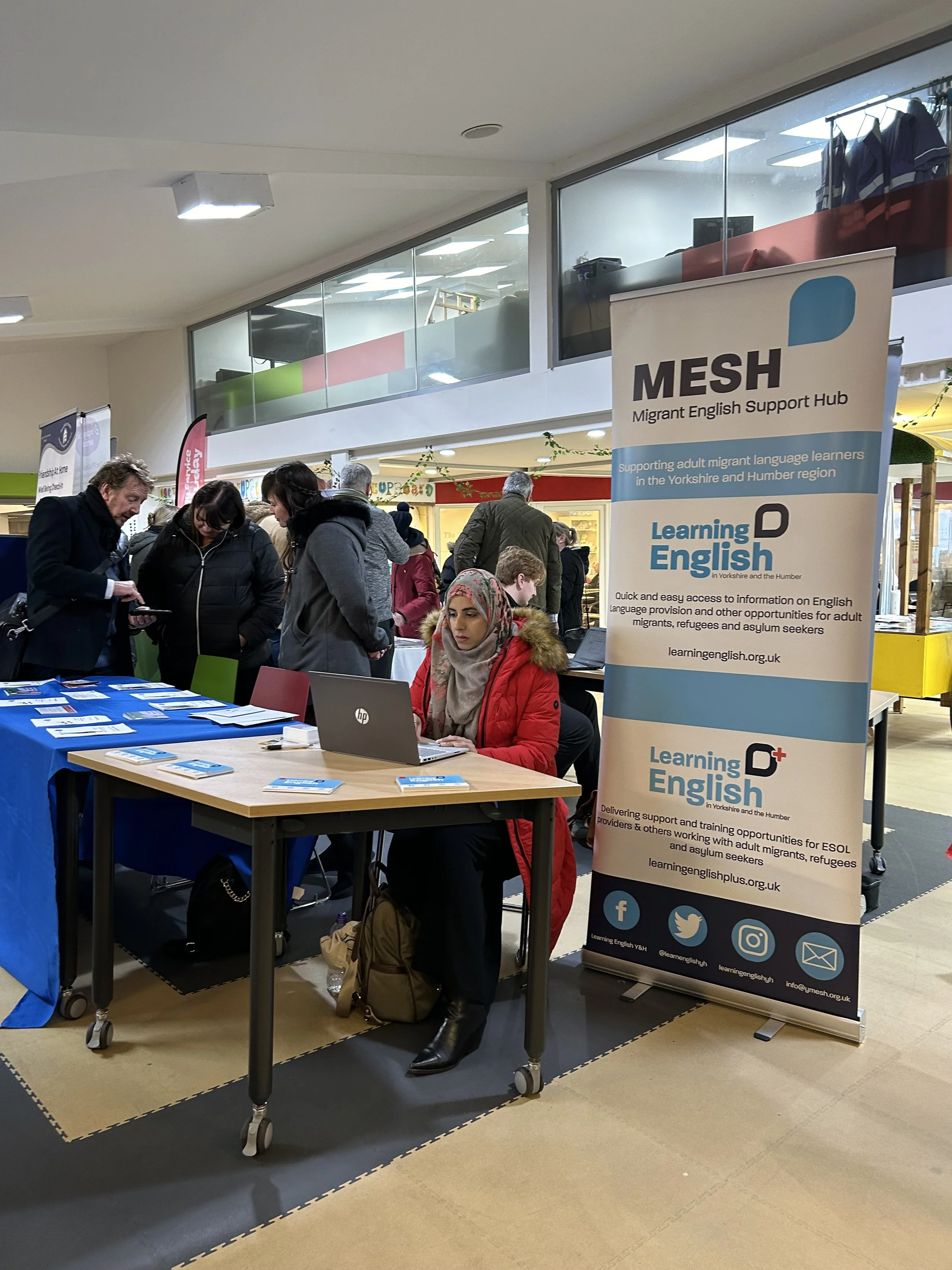 A woman sitting at a table with a laptop and information brochures, at a migrant support event called MESH for English language learners in Yorkshire and Humber, with a large banner behind her displaying the organization's services.
