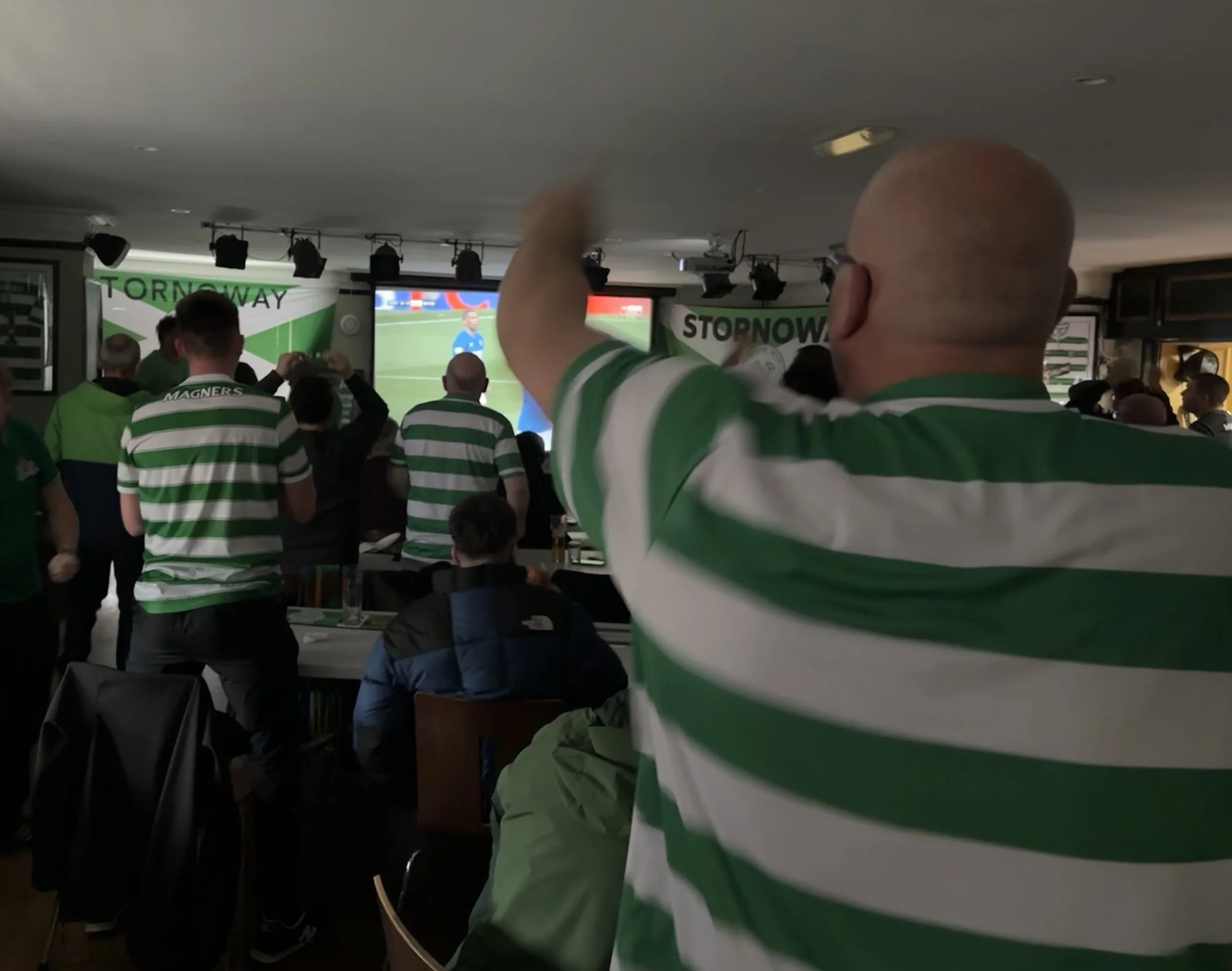 Group of soccer fans wearing green and white striped jerseys watching a game on screens in a bar, with banners reading 'STOPNOWAY' in the background.