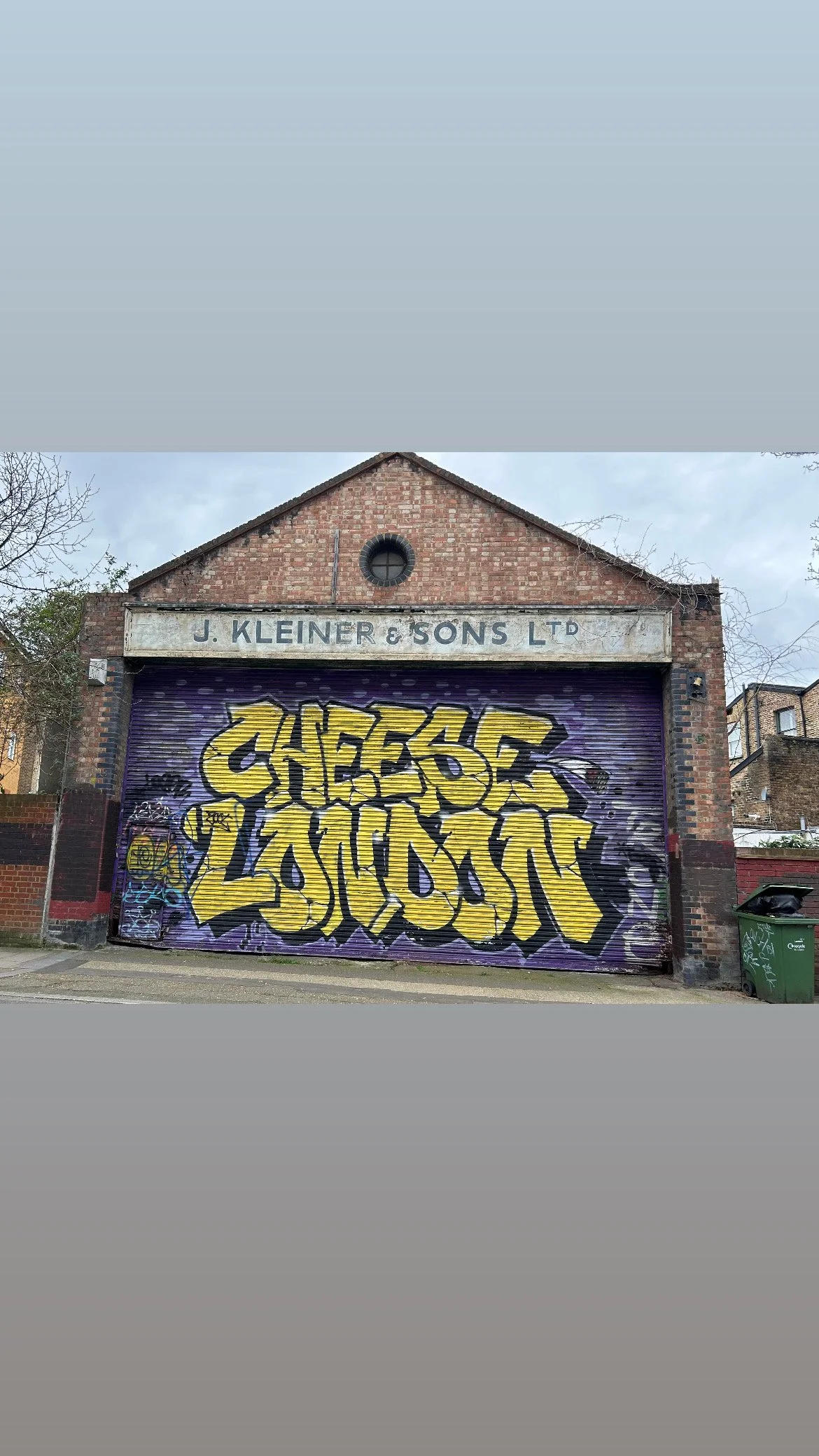 Graffiti art in yellow and black on a purple background with the words "CHEERS TO GOOD" painted on a metal shutter in front of an old brick building labeled "J. KLEINER & SONS LTD."