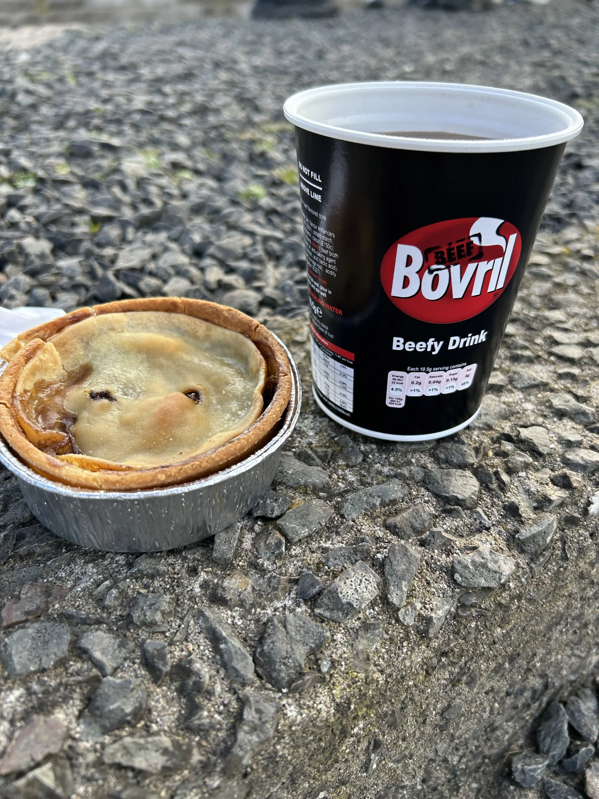 A small aluminum tray with a cheese and meat pie, next to a black cup labeled 'Beef Bovril' on a gravel surface.