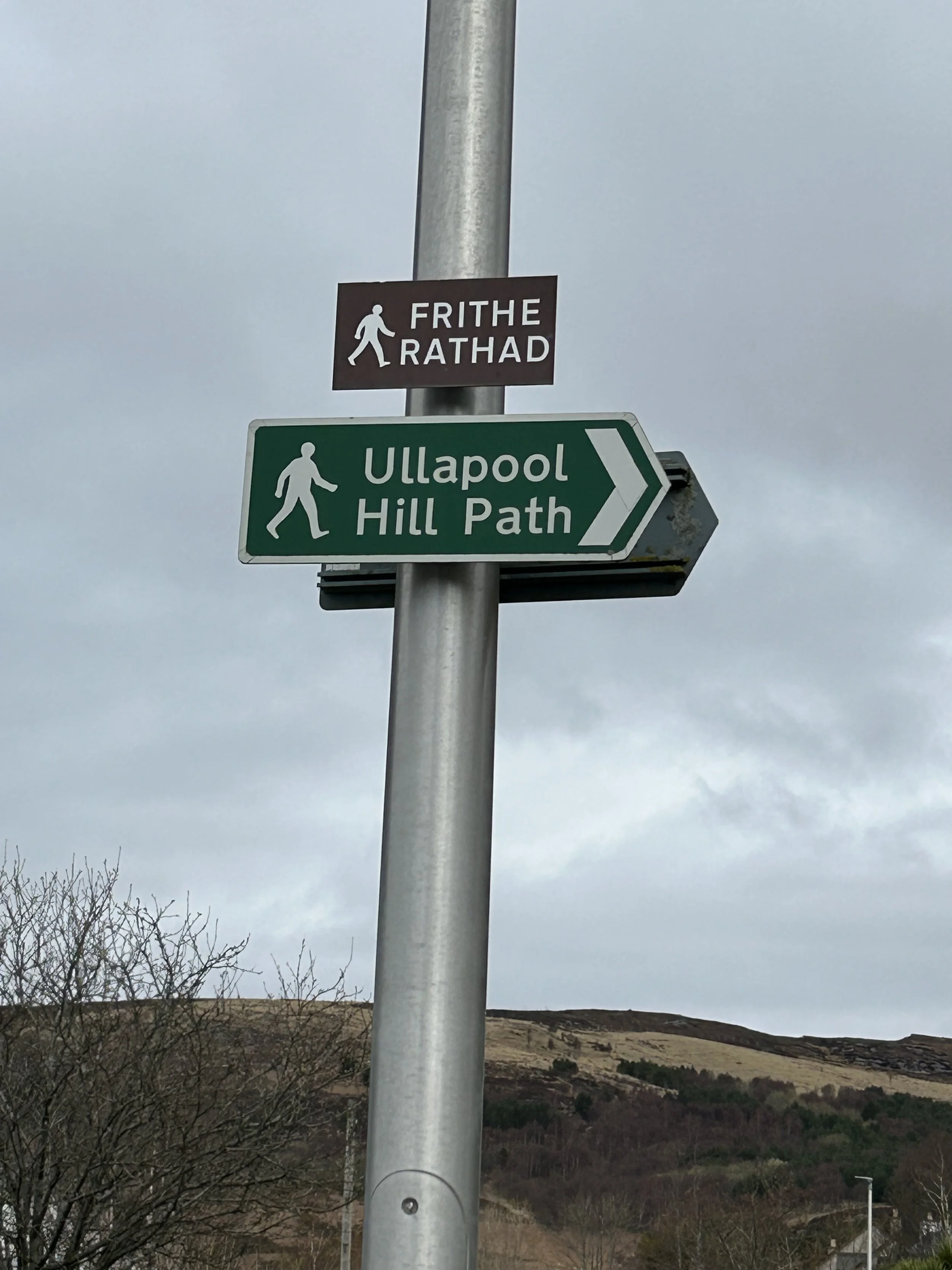 Street signs indicating walking paths, with one sign in Irish (Frithe Radhad) and another in English (Ullapool Hill Path), on a cloudy day with hills in the background.