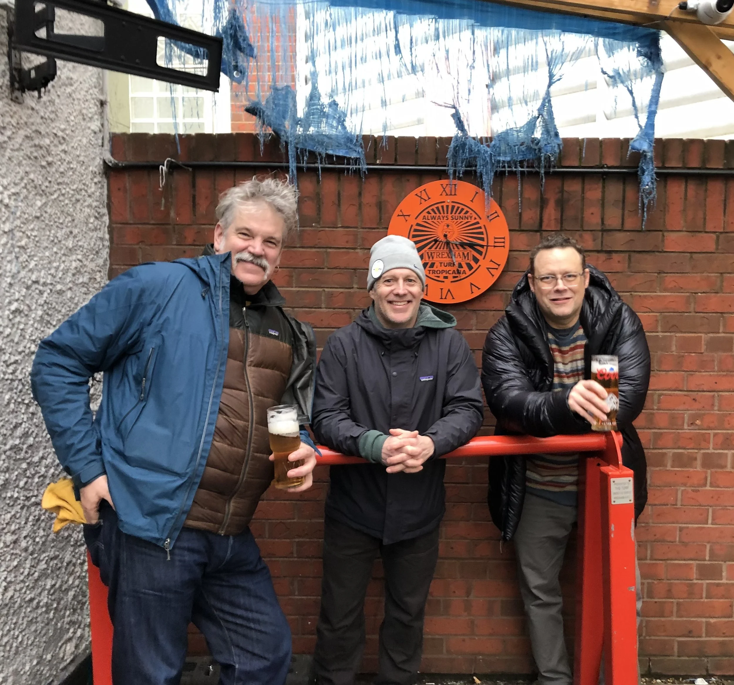 Three men standing outdoors in front of a brick wall, smiling and holding glasses of beer, with a decorative clock and a blue fabric hanging overhead.