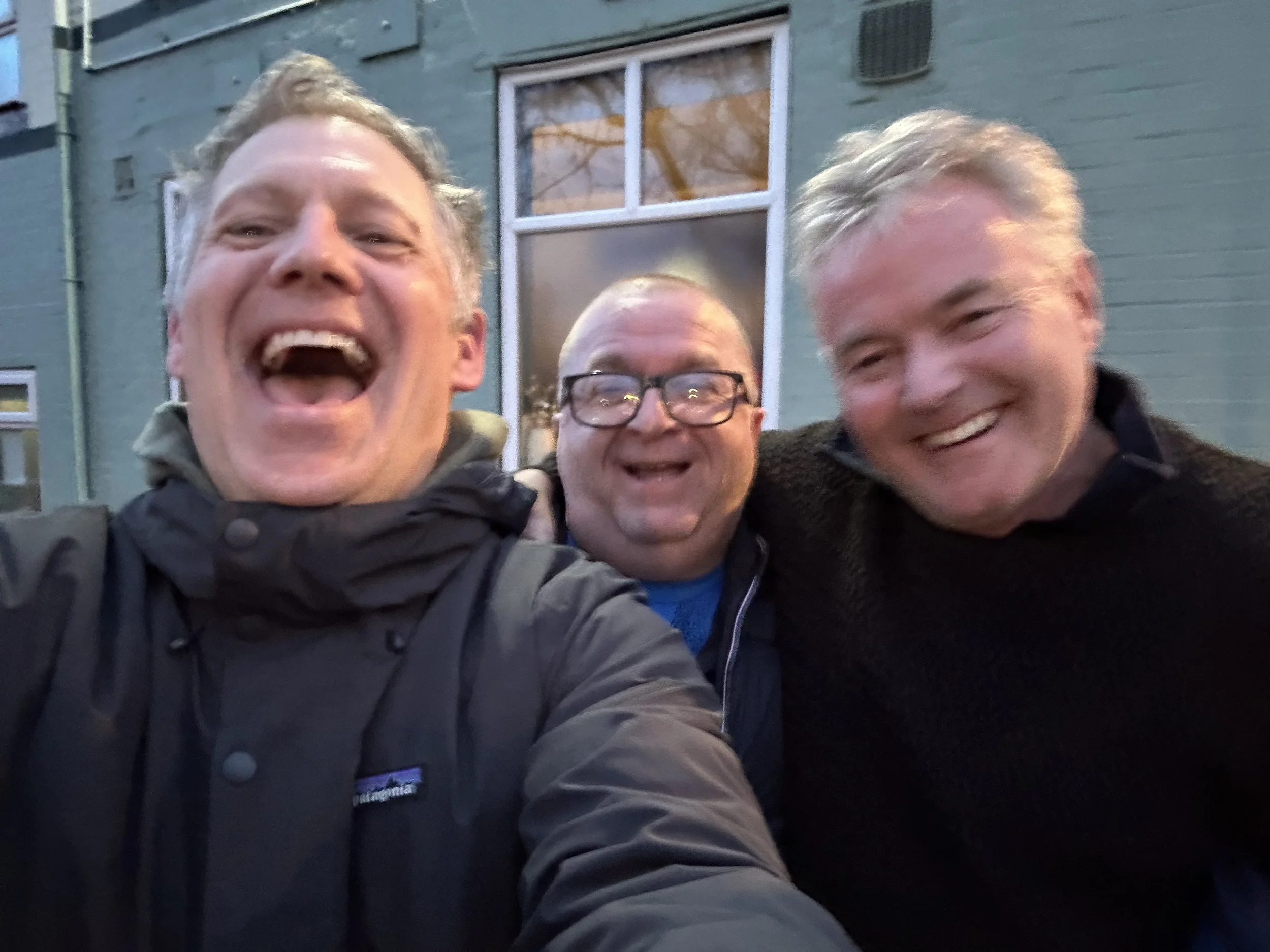 Three men cheerful and smiling taking a photo outside in front of a green building with large windows.