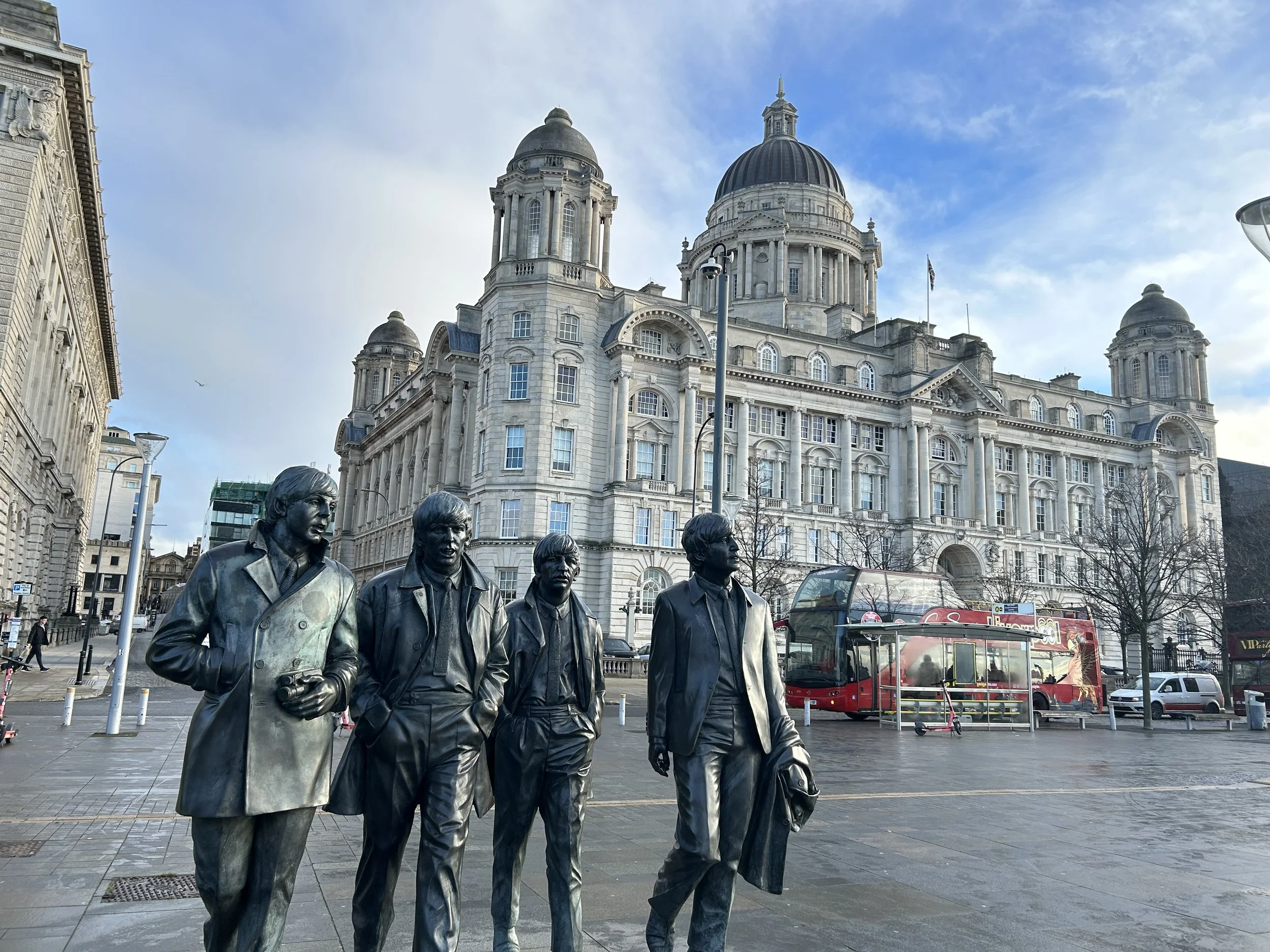 Statue of The Beatles, four men standing on a city sidewalk in front of a historic building with domes and columns, a double-decker bus and some trees in the background.