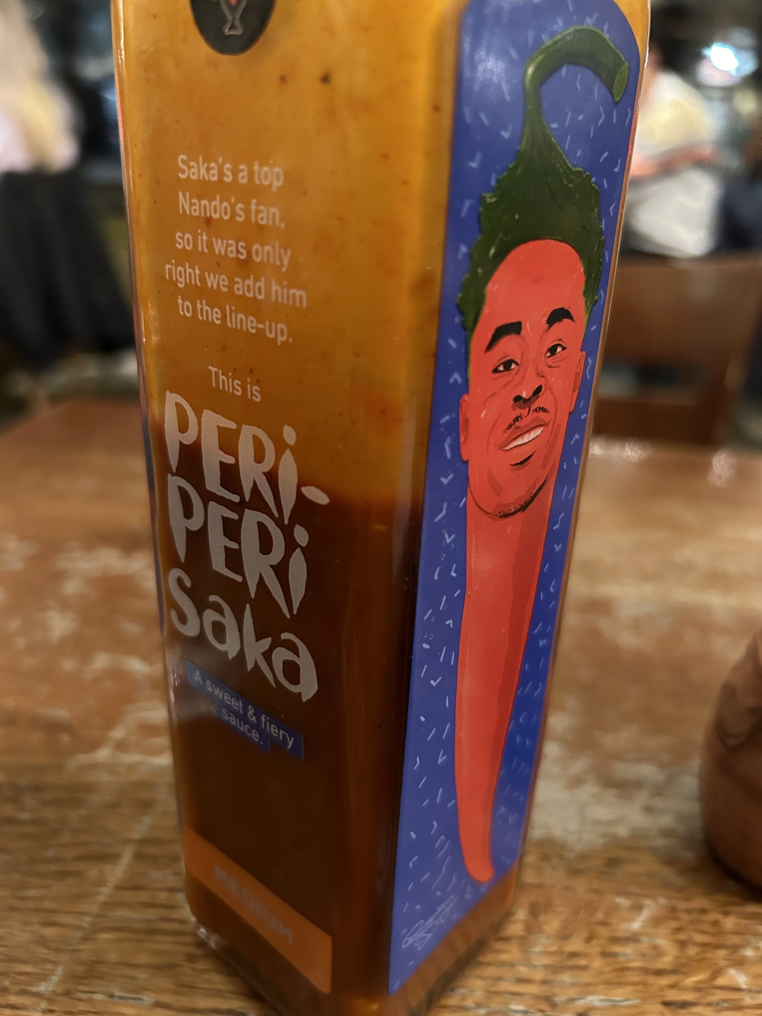 A bottle of Peri-Peri Saka hot sauce with a colorful illustrated label featuring a smiling person's face and a green chili pepper on their head.