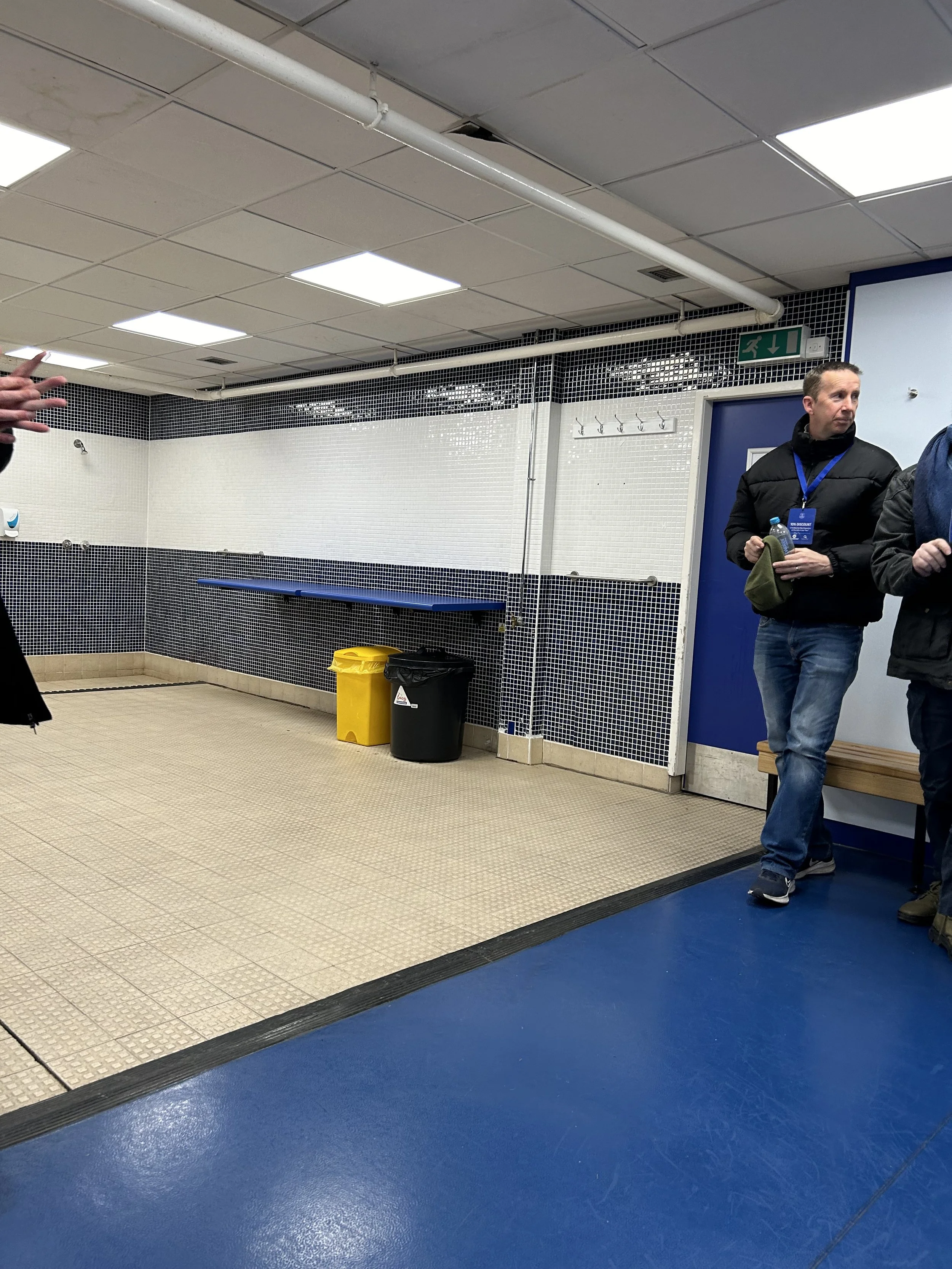 Indoor public space, possibly a hallway or waiting area, with beige and blue flooring, black and white tiled walls, overhead fluorescent lighting, and two people standing near a blue door on the right side of the image.