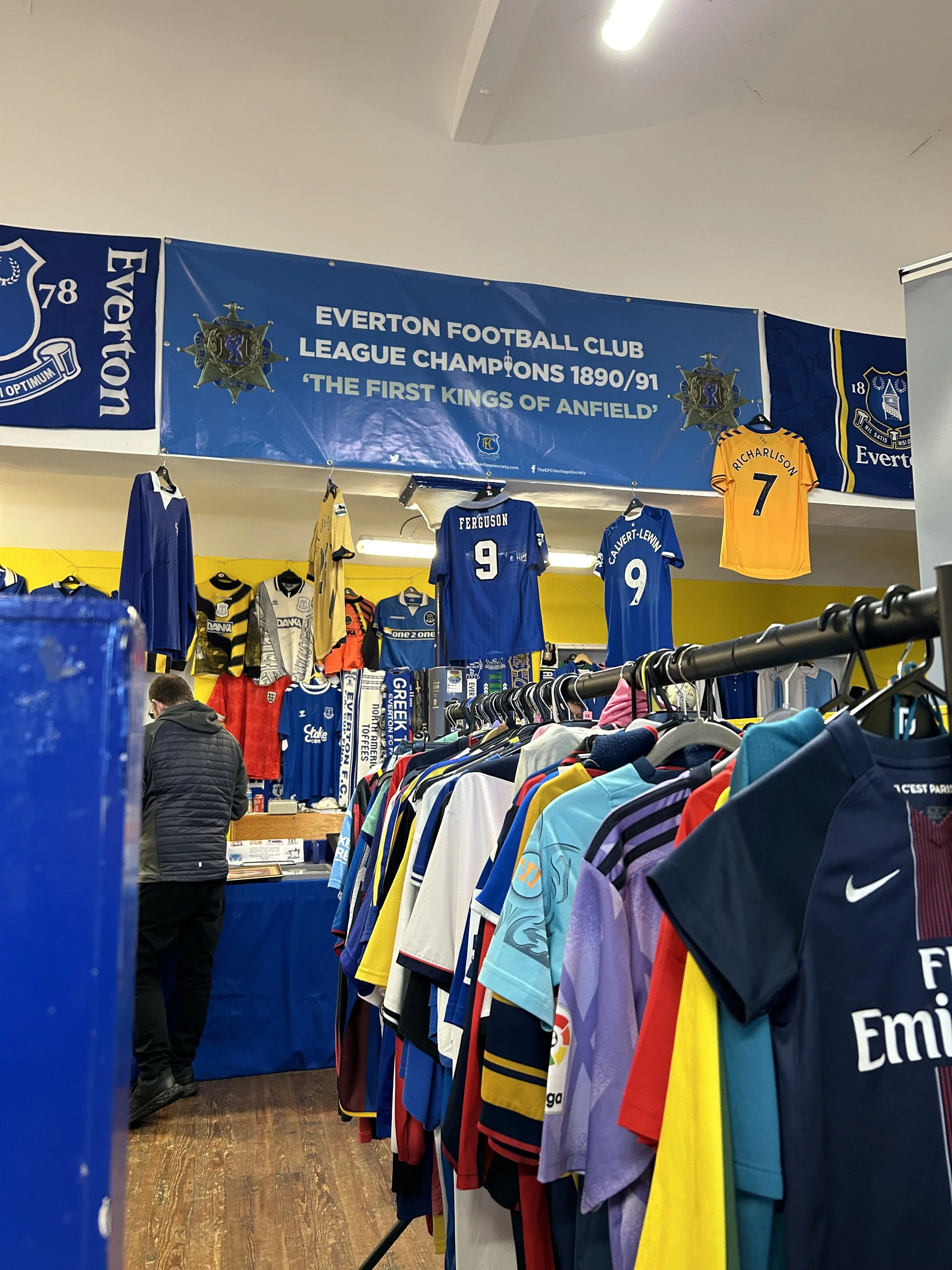 Inside a store selling Everton Football Club merchandise, with jerseys, flags, and shirts displayed on racks and walls, including banners commemorating the club's league championship in 1890/91.