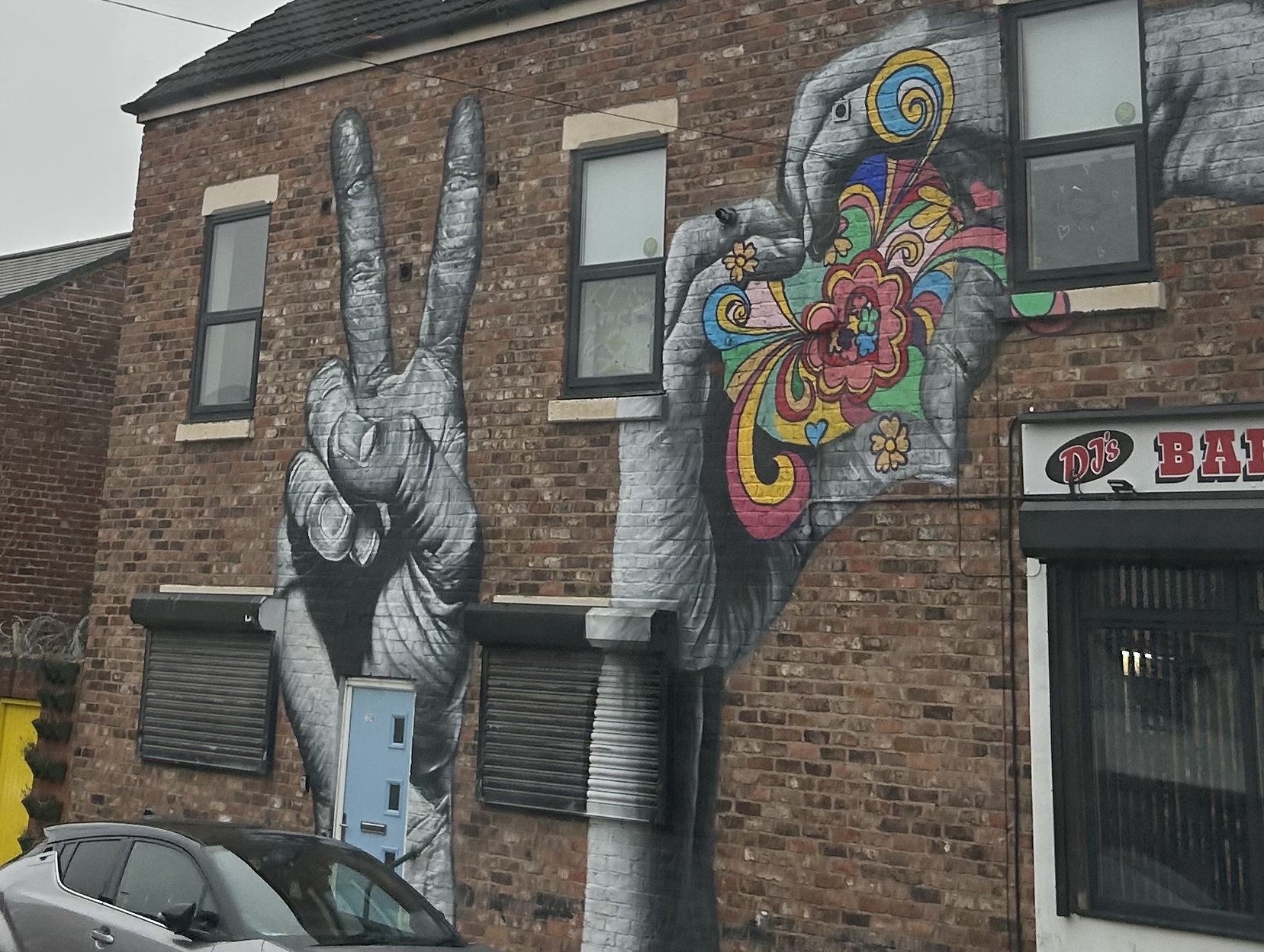 A mural on a brick building showing two hands, one making a peace sign and the other holding a colorful, floral pipe.