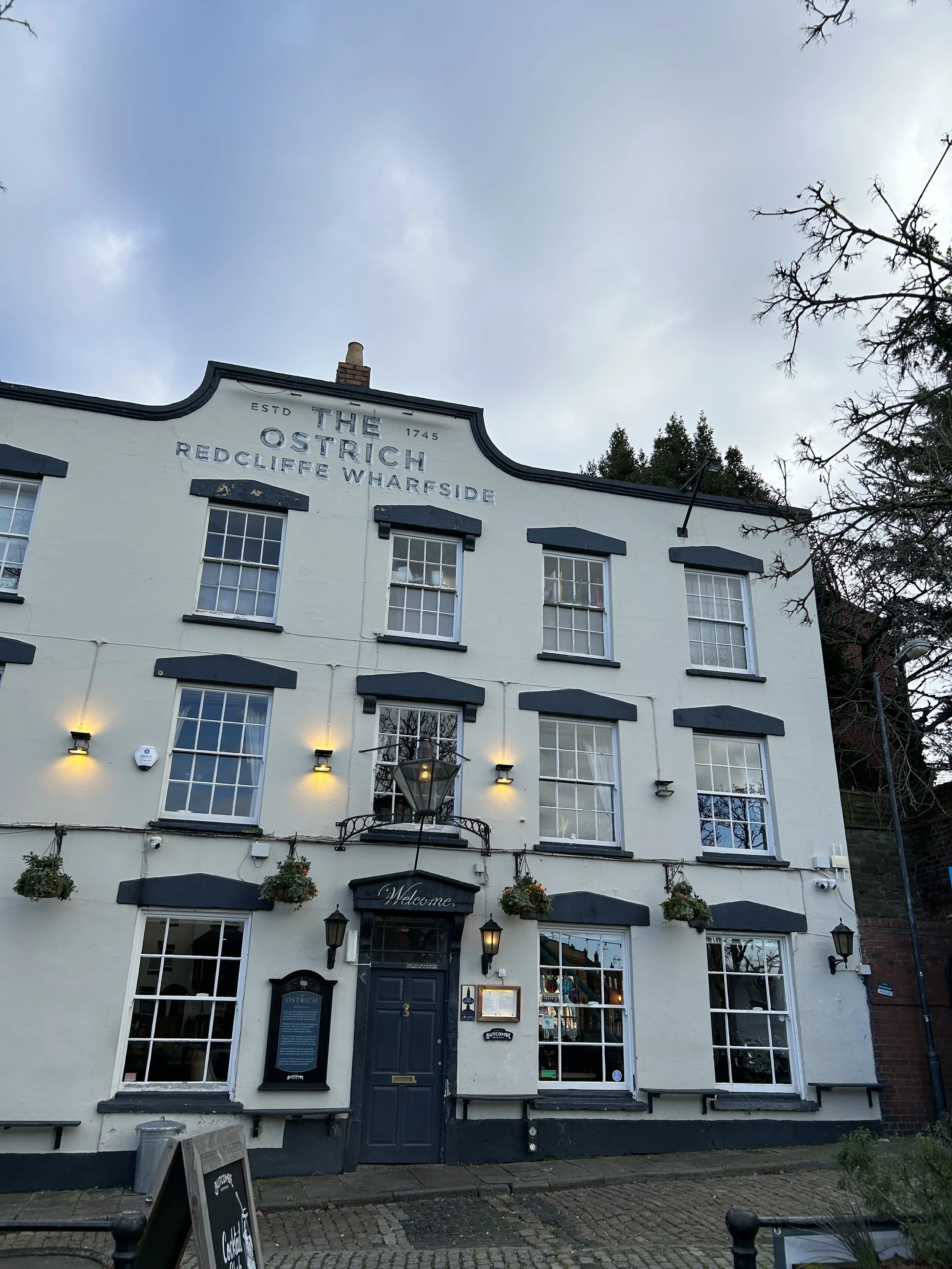 A four-story white building with black accents and multiple windows, labeled 'The Ostrich Redcliffe Wharfside,' with a sign reading 'Welcome' above the door, hanging flower baskets, and outdoor lighting. Overcast sky with trees on the side.