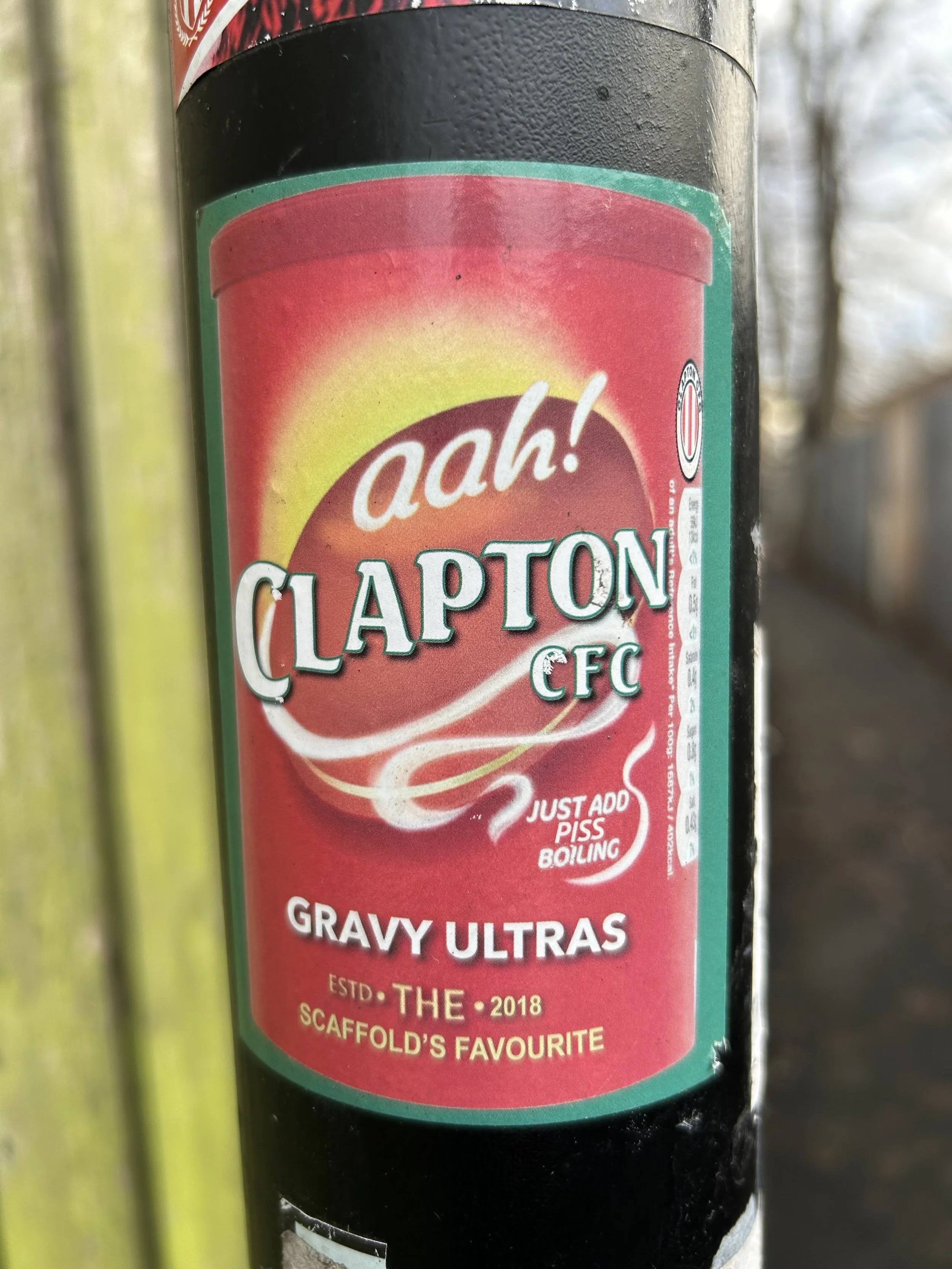 Close-up of a black post or pole with a humorous parody sticker resembling a fast food sauce label, featuring red, yellow, and green colors, the word "aah!" in white script, and the phrase "Clapton CFC" with additional text about gravy and a humorous