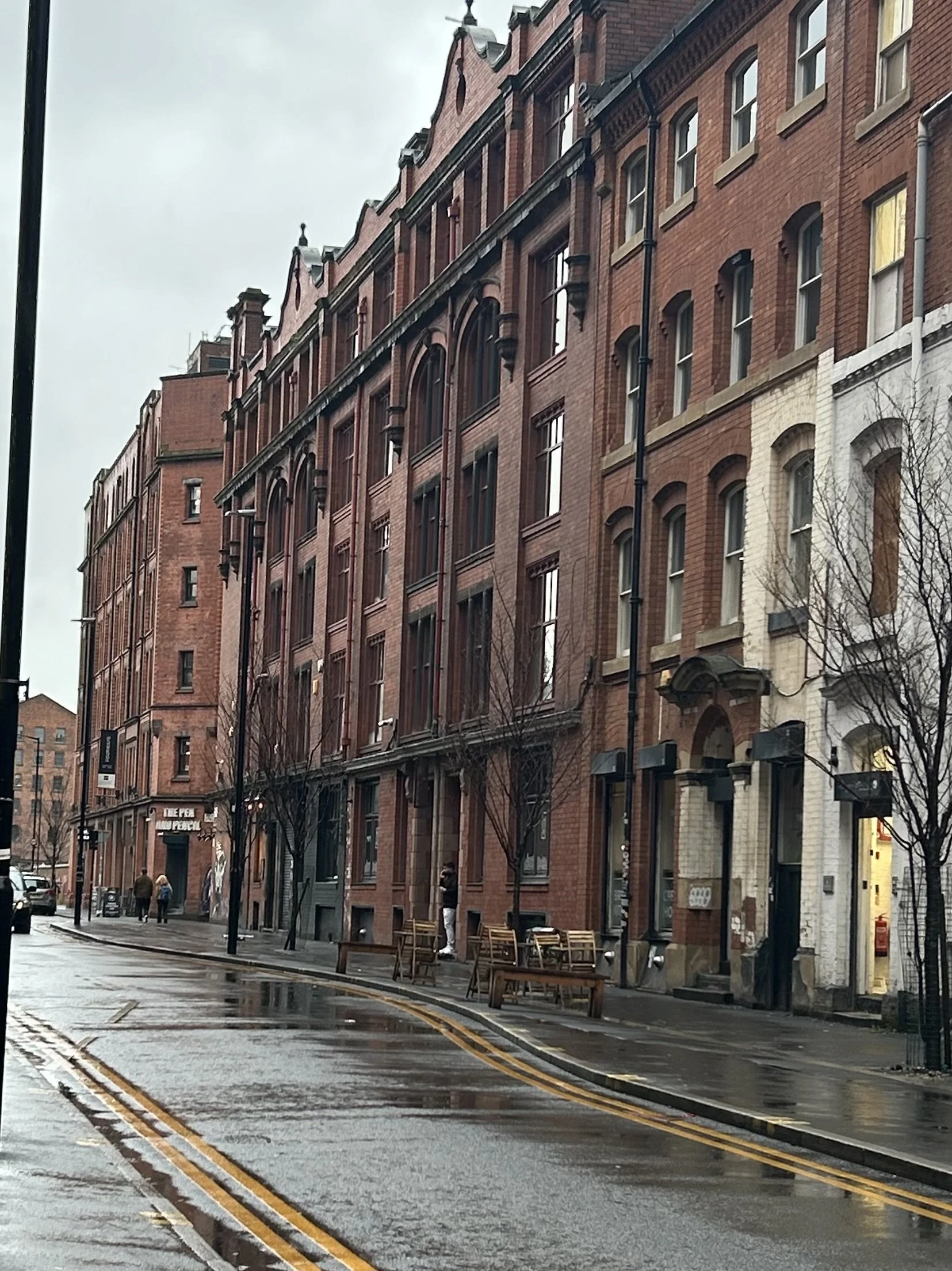 Rain-soaked street lined with red brick buildings, some with arched windows, and benches along the sidewalk. A few pedestrians walk in the distance on a cloudy day.