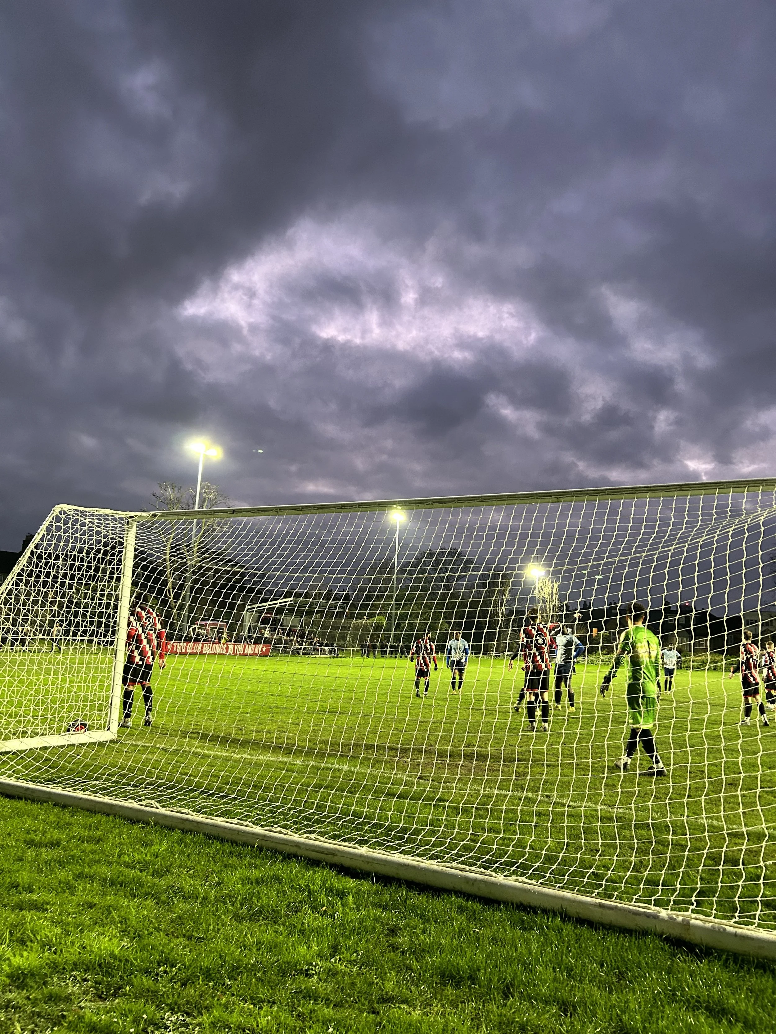 A soccer field with players and a goalkeeper under a dark, cloudy sky, illuminated by stadium lights.