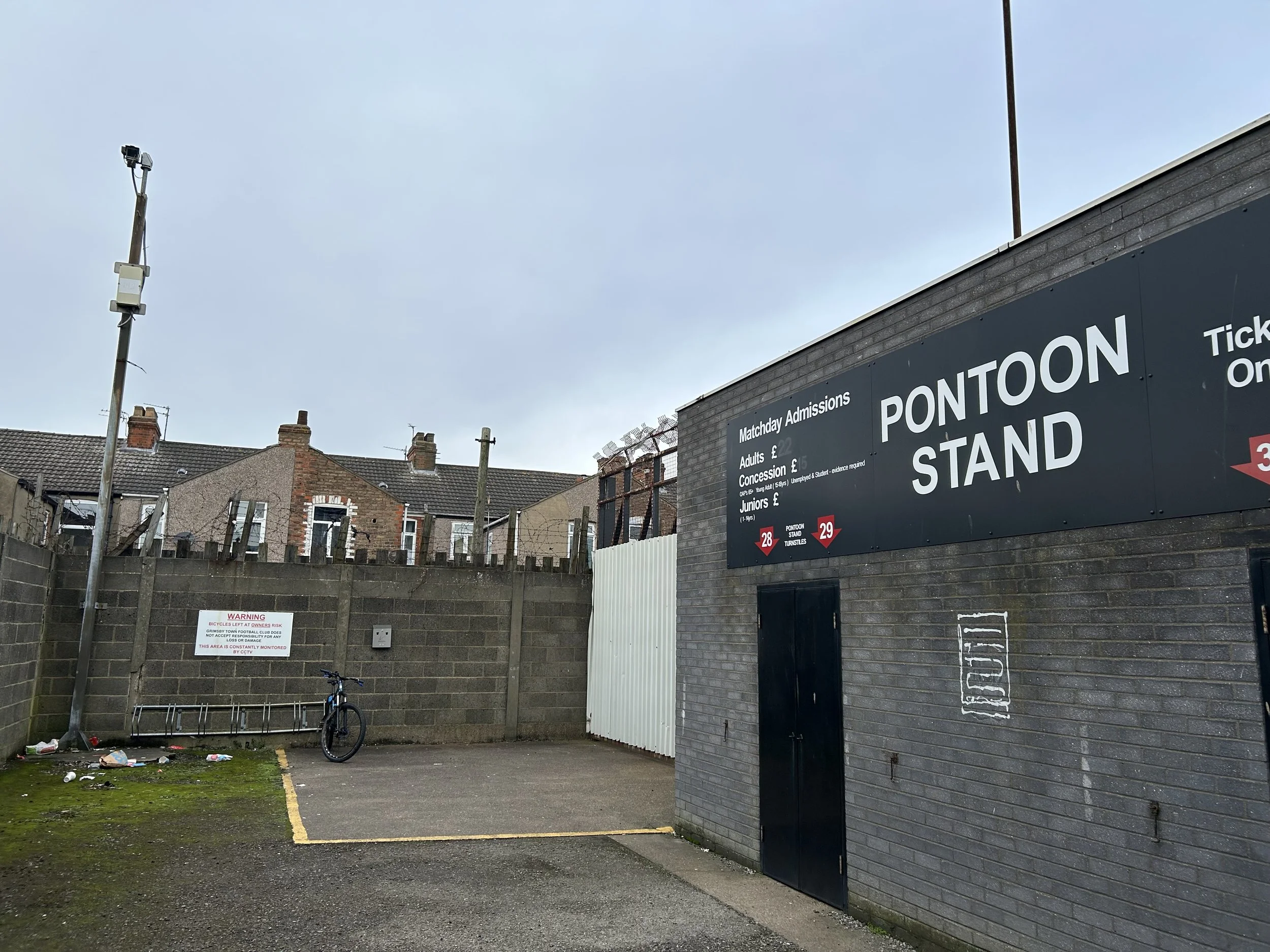 A parking lot next to a building with a large sign that reads "PONTON STAND". The sign also lists ticket prices for matchday admissions, including adults, concession, and juniors. A bicycle is leaning against a concrete wall, and there is litter on t