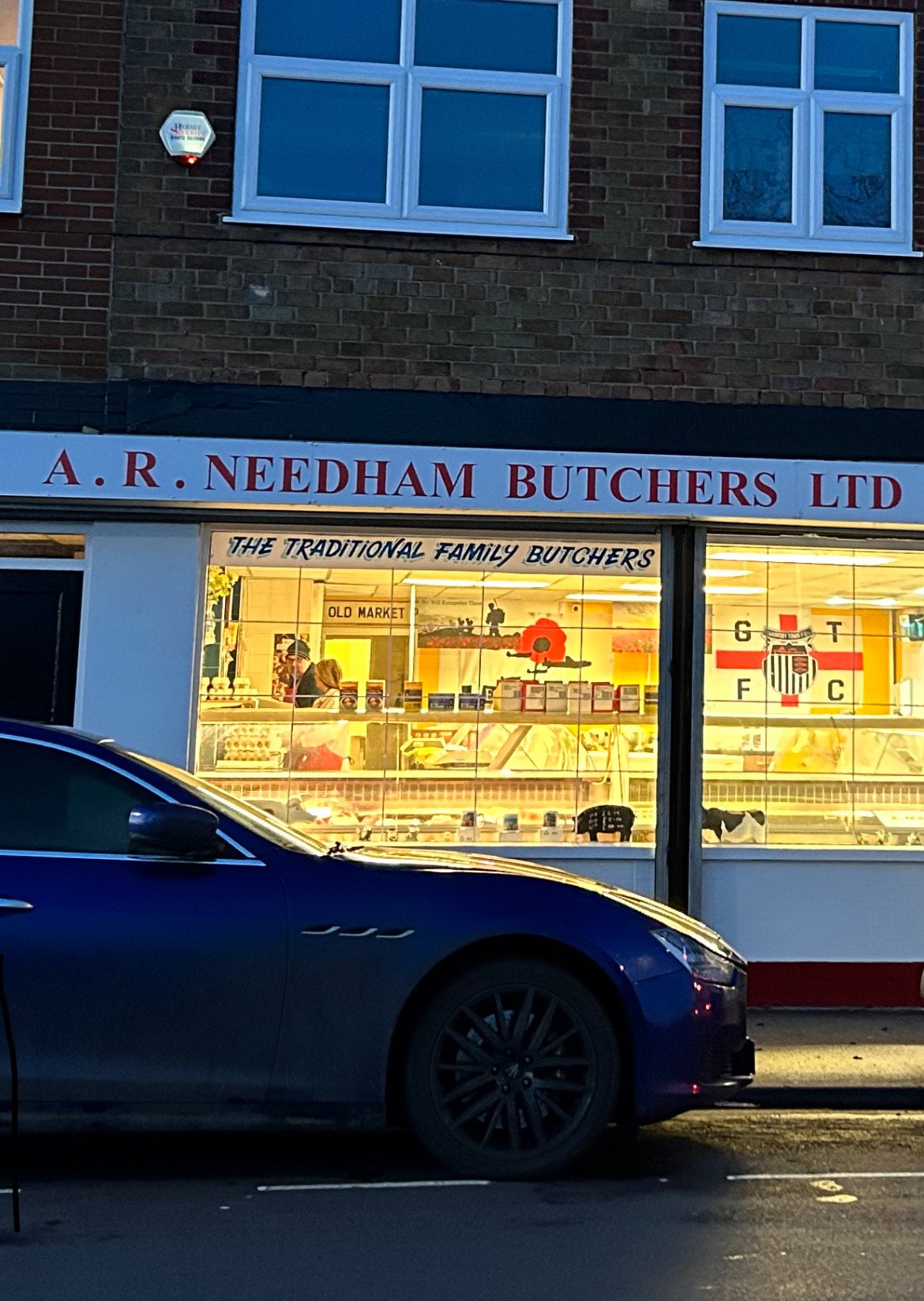 A butcher shop named A. R. Needham Butchers Ltd with a yellow illuminated display window showing meats and people inside. The storefront has a sign with the butcher's name and a slogan "The Traditional Family Butchers." In front of the shop, a blue c