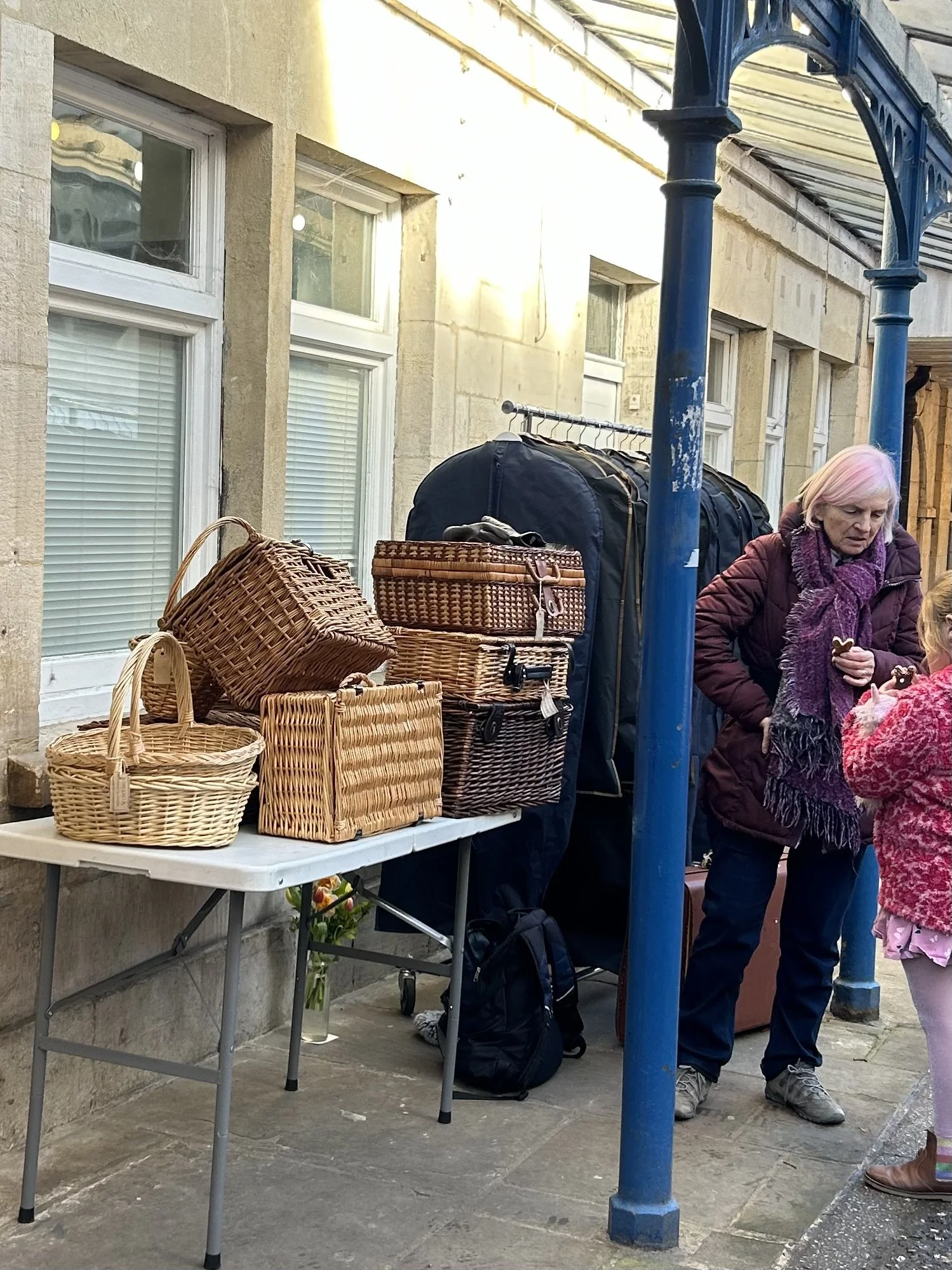 Street market stall with wicker baskets on a table, a large bag, backpacks, and a woman with pink hair talking to a child, under a blue metal canopy.