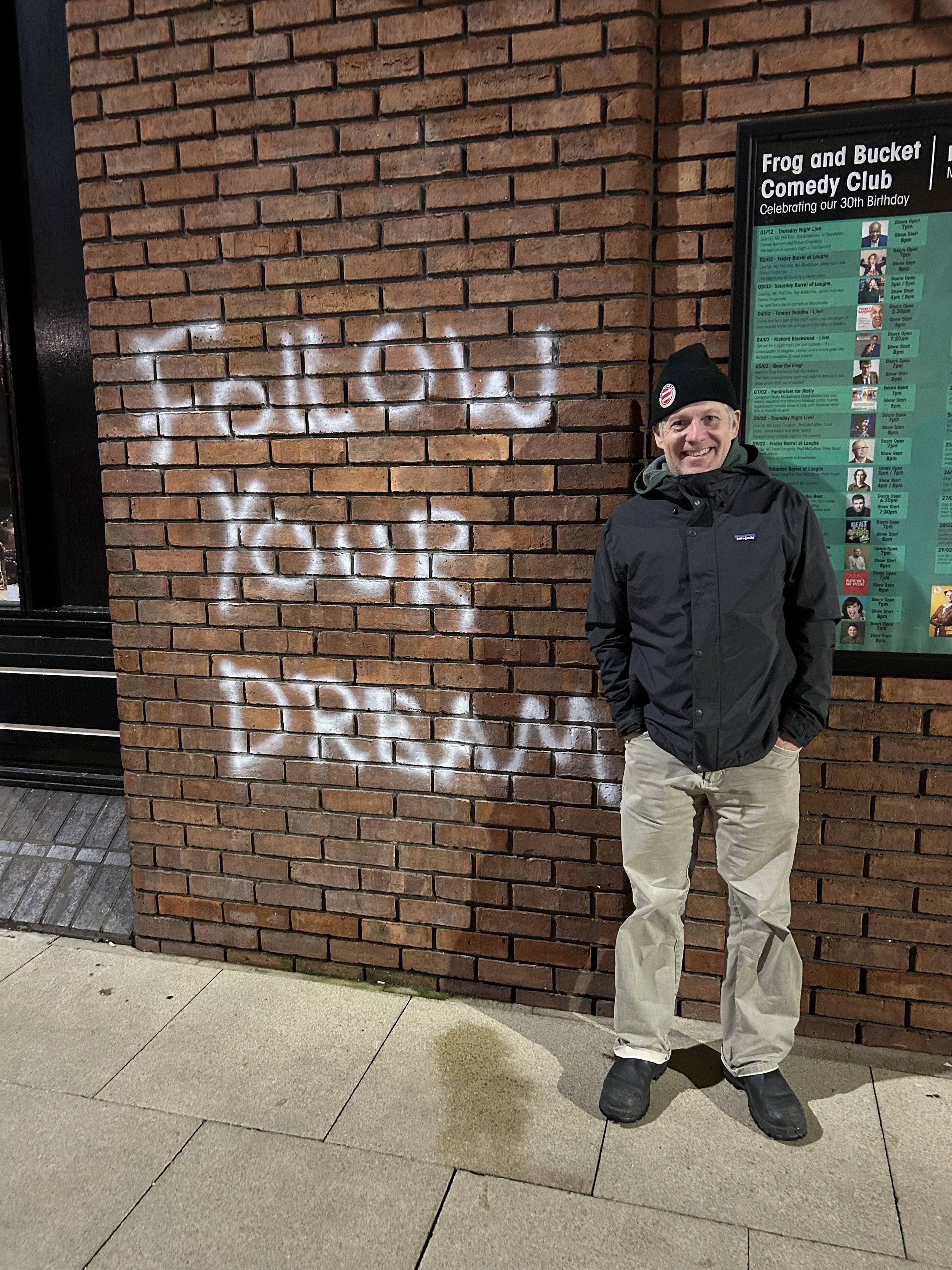 A man in a black jacket, beige pants, and black beanie standing outside next to a brick wall with white graffiti. There is a green and black podium behind him with a schedule for a comedy club event.