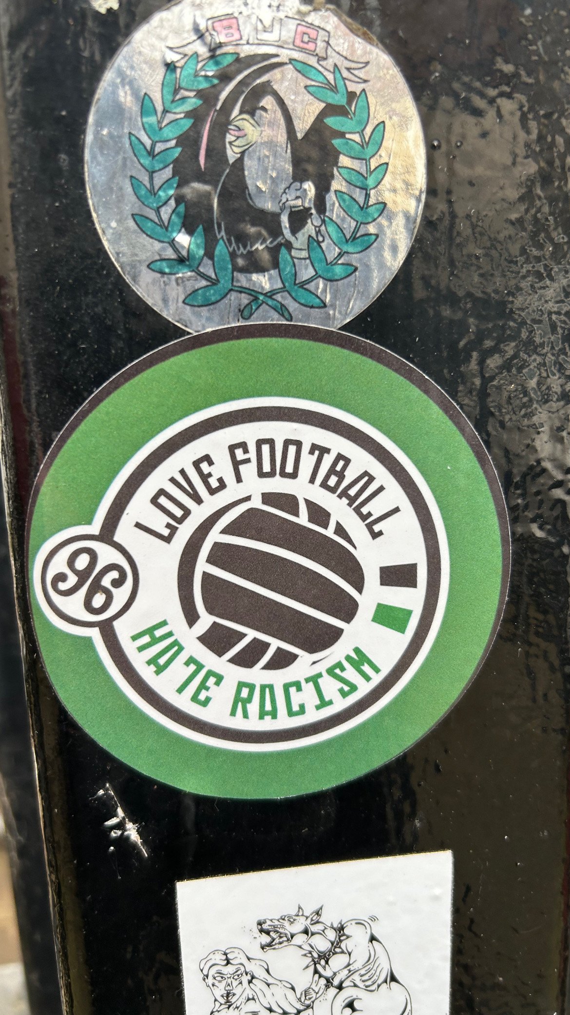 Stickers on a black surface, including one with a graphic of a bald eagle, a volleyball with text 'Love Football Hate Racism,' and a line art drawing of a young man and a wolf.