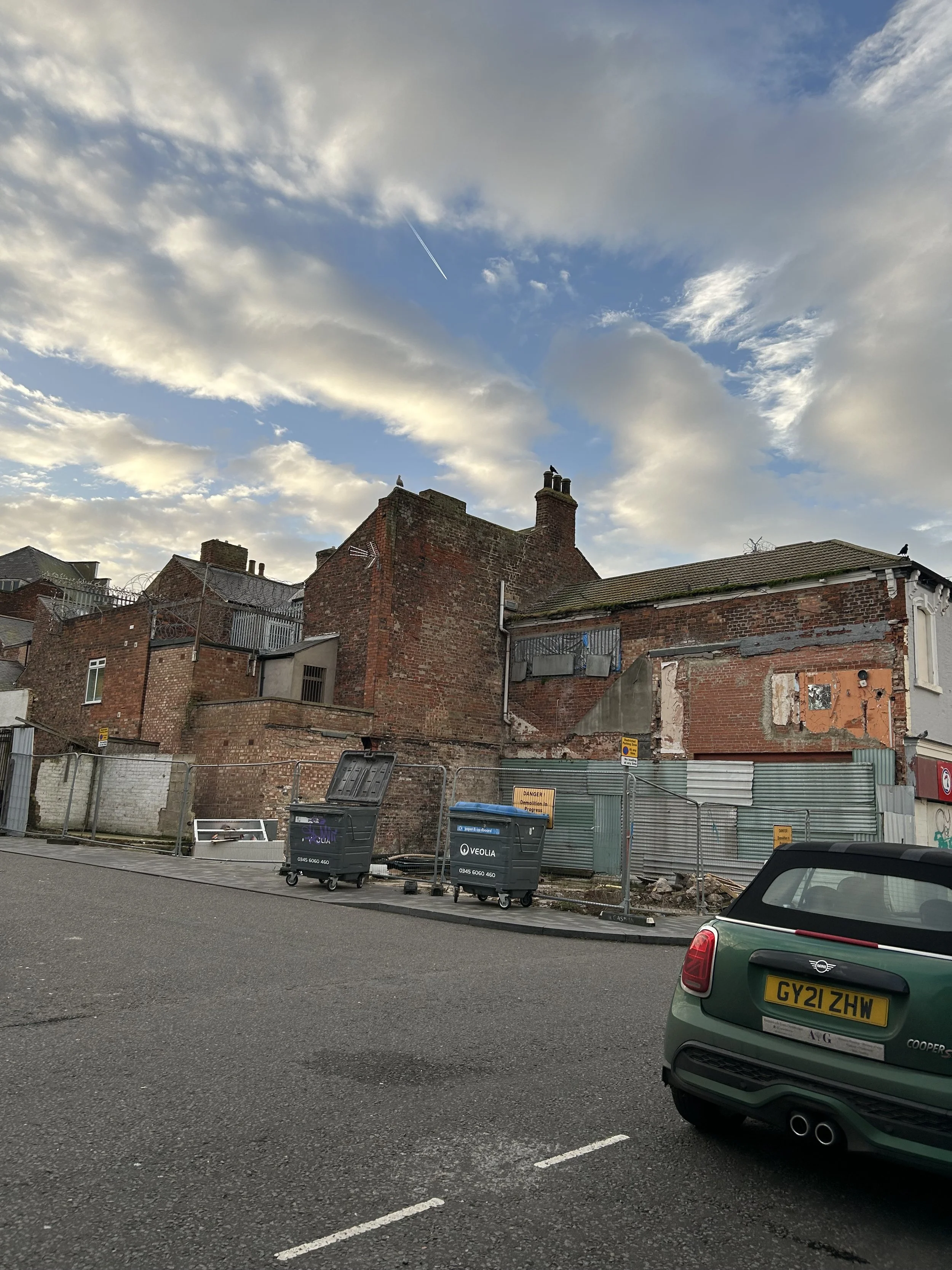 An urban scene featuring a partly demolished brick building with construction fencing, trash bins, a green Mini Cooper car with a UK license plate, and a sky with clouds and a jet stream.