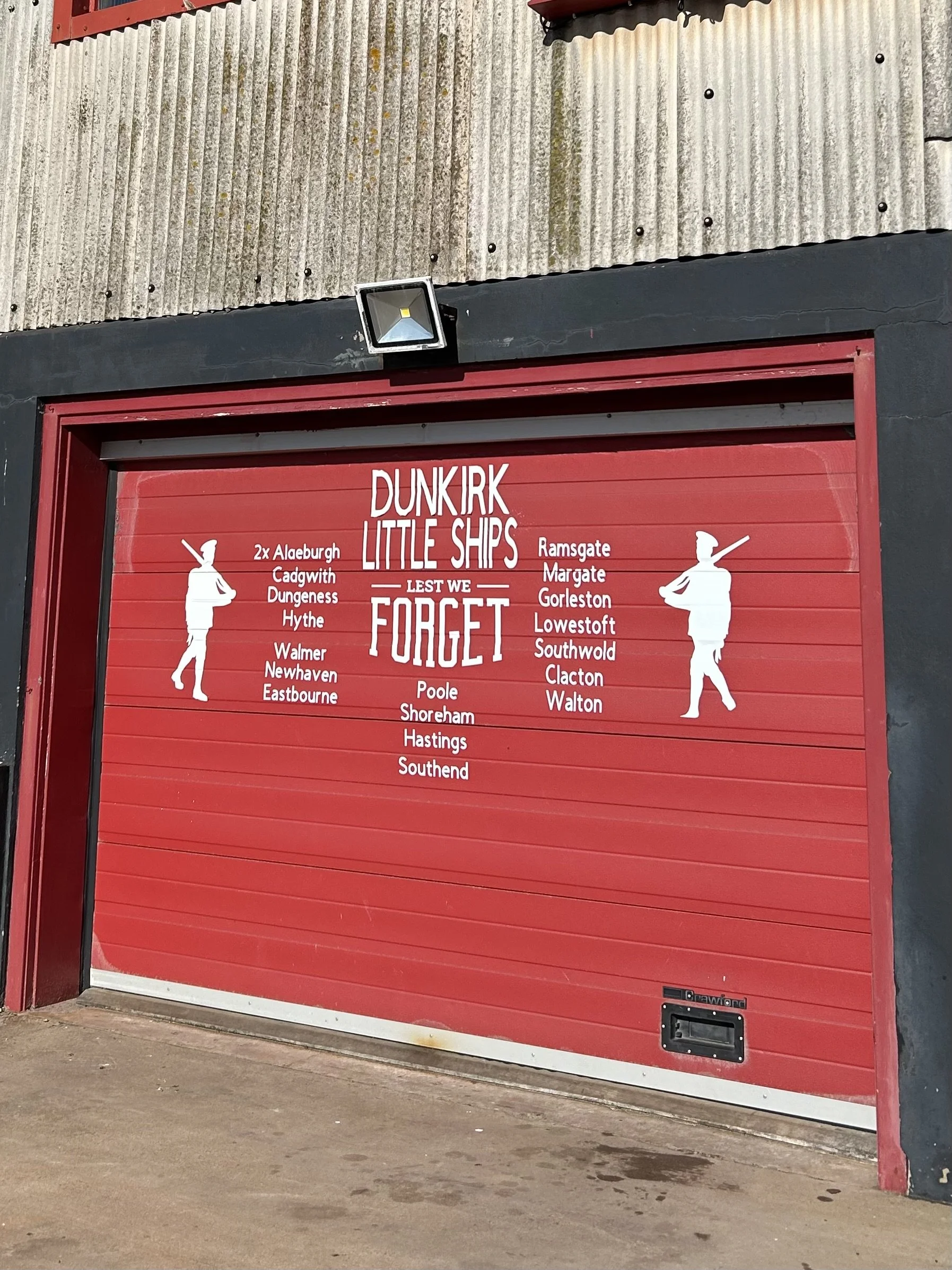 Red garage door with a sign titled 'Dunkirk Little Ships' displaying lists of ship names on each side, with two white silhouette illustrations of sailors or naval personnel on each side.