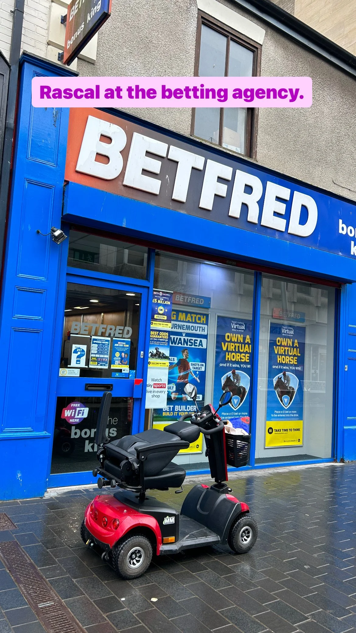 A red mobility scooter parked outside a blue betting shop named Betfred. The shop has promotional posters and signs in the window, including a sign that says 'OWN A VIRTUAL HORSE.' The sidewalk is wet, and there is a building with a window above the 