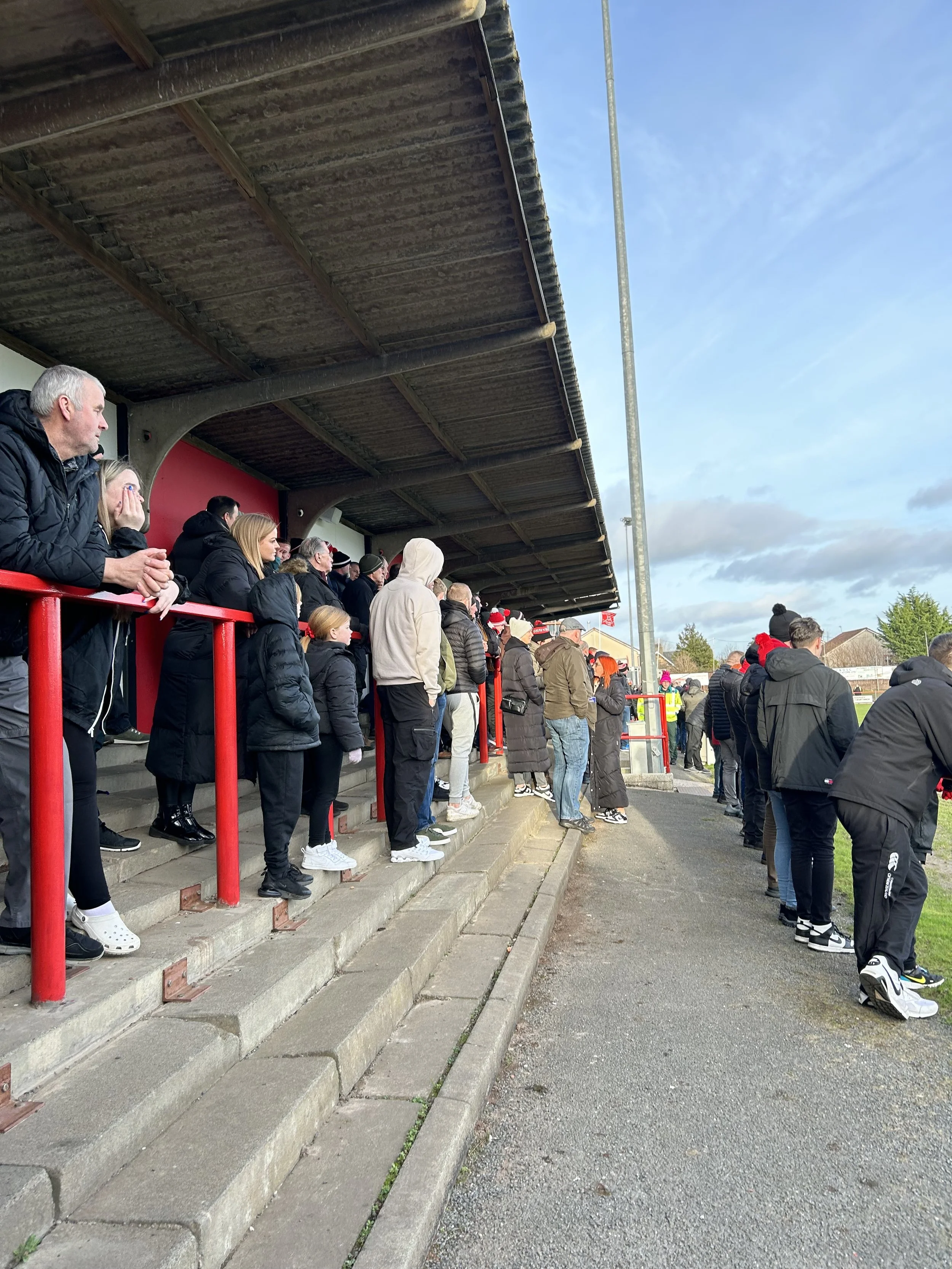 Crowd of people standing on steps at a baseball game, wearing winter clothing, with some watching the game on the field, under a covered seating area.