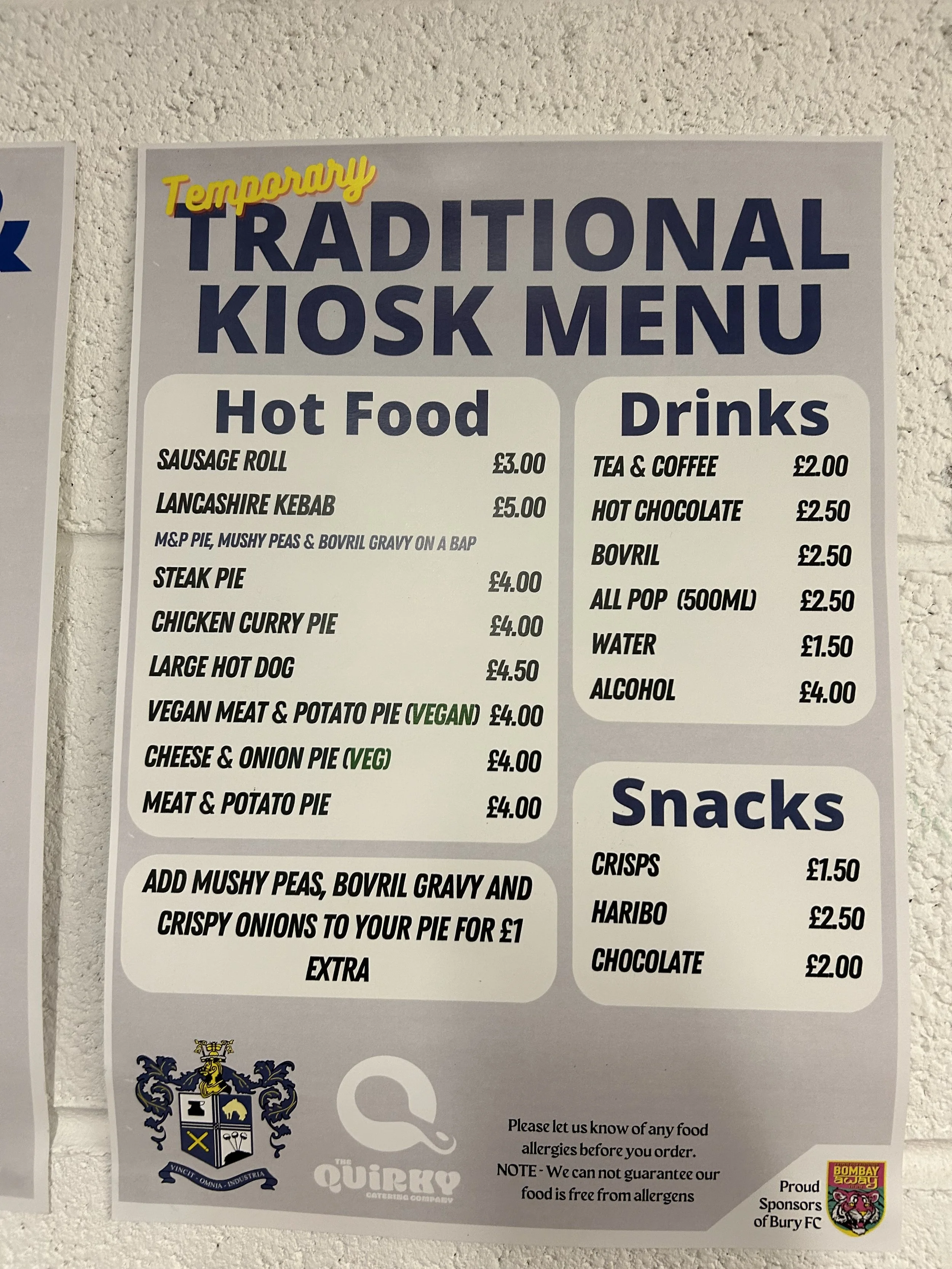 A menu board listing various hot foods, drinks, and snacks at a temporary kiosk. The hot foods include sausage roll, Lancashire kebab, steak pie, chicken curry pie, large hot dog, vegan meat and potato pie, cheese and onion pie, and meat and potato p
